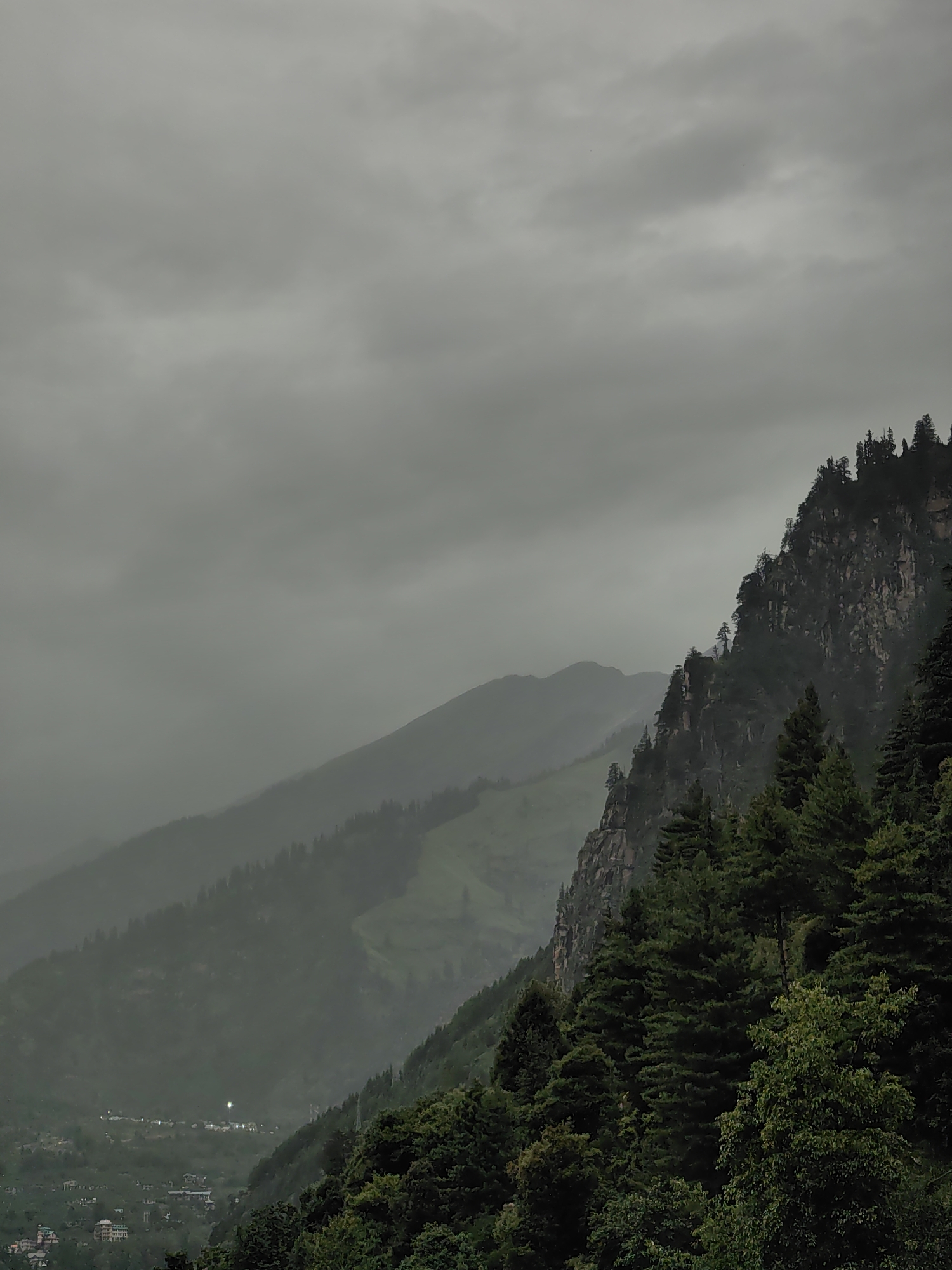 The image depicts a mountainous landscape under a cloudy sky. The foreground features a dense forest of dark green coniferous trees, with some deciduous trees interspersed. A steep, rocky cliff face rises behind the trees on the right side of the image. Further in the background, several layers of mountains recede into a misty haze. In the valley between the mountains, a small settlement is visible, with clusters of buildings and lights indicating a town or village. The overall atmosphere is somber and moody, consistent with overcast weather. Based on the distinctive terrain and the architectural style of the buildings in the settlement, this scene is likely located in or around Vashisht, India, a town known for its mountainous setting and natural hot springs. There are no people or discernible actions depicted in the image.
