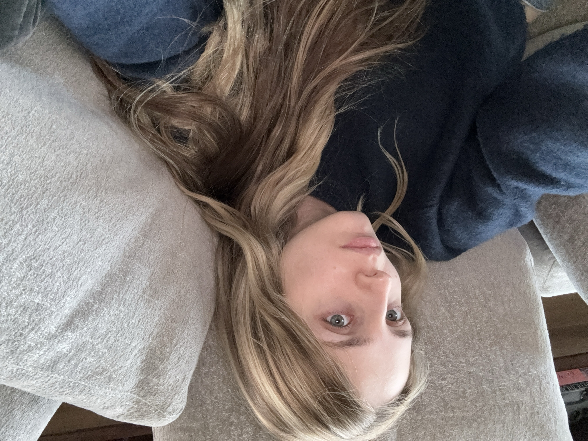 A woman with blonde and brown hair is reclining on a gray textured sofa, wearing a dark blue sweater. She is looking directly at the camera with pale skin and light-colored eyes. Her head is resting on a light gray pillow or the arm of the sofa. The image is taken from above, with the perspective rotated counter-clockwise. In the lower right corner, there are some books with writing on their spines. The location is not identifiable as Mexico City from the details in the image.