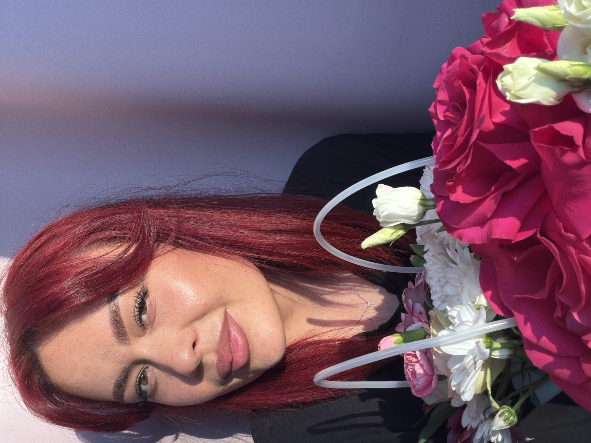 A close-up shot shows a young woman with bright red hair and freckles, smiling gently as she holds a bouquet of flowers. She is wearing a black top and a delicate silver necklace. The bouquet contains a variety of flowers, including large pink roses, white chrysanthemums, and pink carnations. The background is a plain, light purple wall, with a shadow cast from the woman's left side. The lighting suggests it is daytime, with sunlight hitting the woman's face from the right. The image is framed from the chest up, focusing on the woman's face and the colorful flowers. The location is unspecified in the image itself, but the prompt asks for it to be in the city of Узюково, Russia.