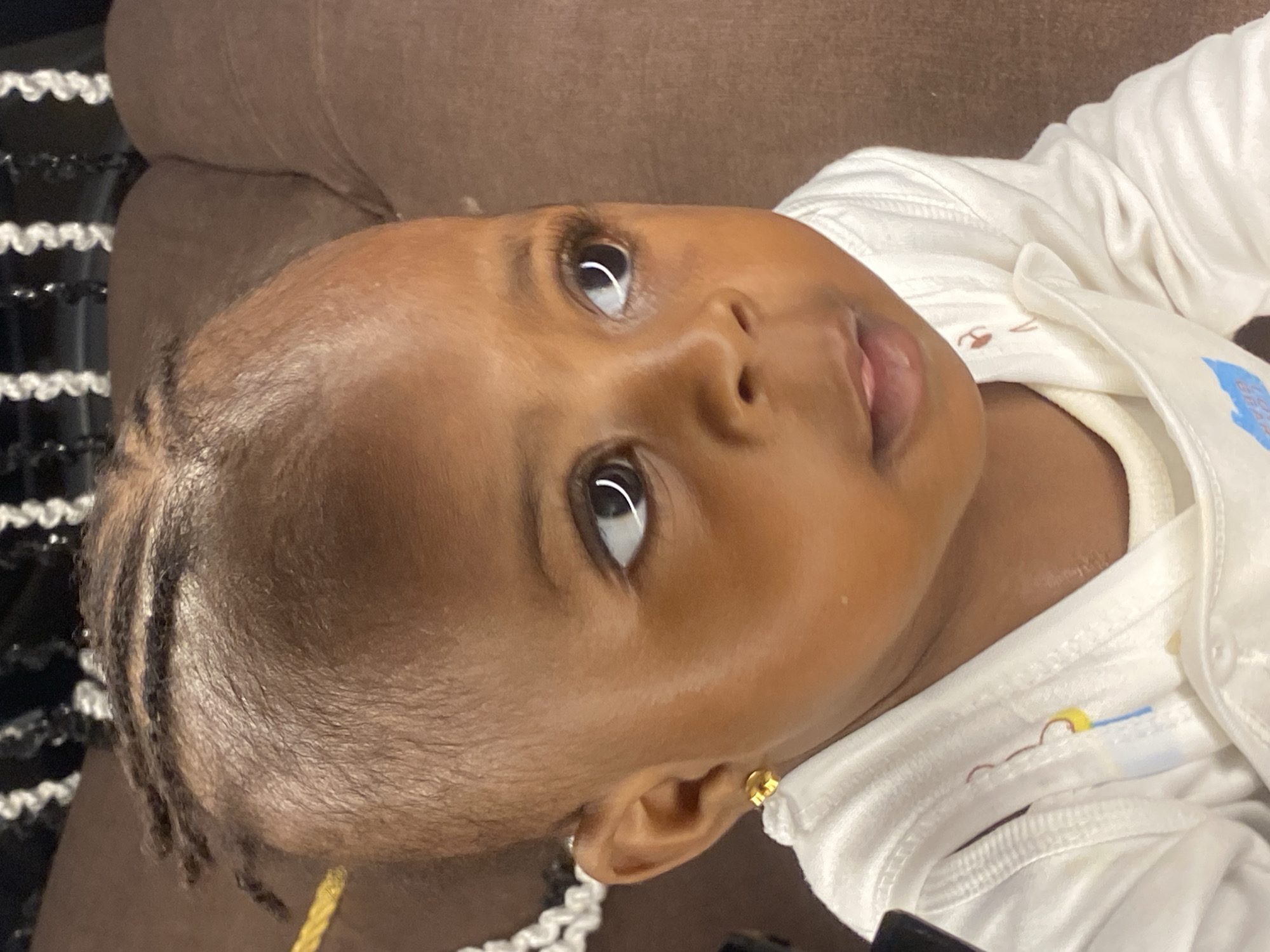 This is a medium shot, eye-level, indoor photograph of a young Black baby girl with dark skin and dark braided hair. She is wearing a white long-sleeved onesie with a few colorful embroidered designs on the chest, including a yellow circle with a blue line and a small brown bear outline. She has a gold stud earring in her left ear. The baby is looking to her right, with her mouth slightly open. Her eyes are wide and dark, with long eyelashes. The background is a blurred brown surface, likely a couch or chair. The lighting is soft and appears to be natural. The image is in focus on the baby's face and upper body.
The specific location in Abakaliki, Nigeria, cannot be determined from the provided image.