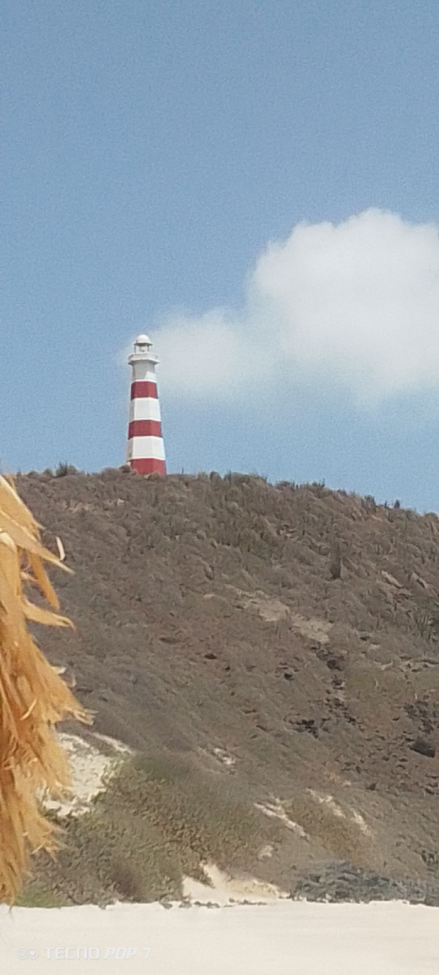 The image depicts a lighthouse situated on a hill in Pedro González, Venezuela. The lighthouse is striped red and white. A light grey cloud is adjacent to the right side of the lighthouse. The hill is covered with sparse vegetation. In the foreground, a light brown object, possibly a thatched umbrella, is partially visible on the left. The sky is blue with scattered clouds. The sand on the beach can be seen at the bottom of the picture.