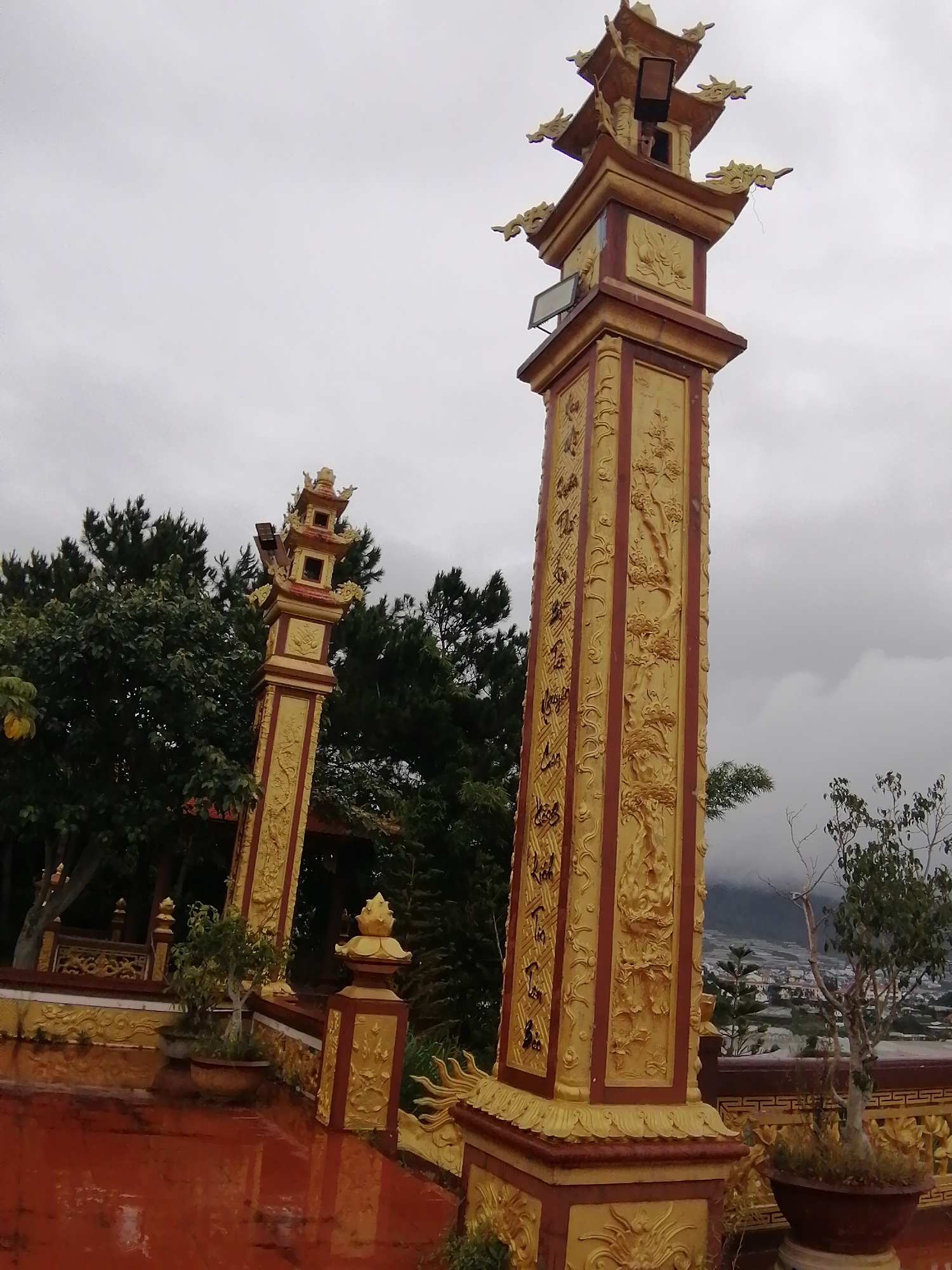 The image depicts an ornate golden pagoda-style pillar with intricate carvings, set against a cloudy sky. A second, similar, but smaller pillar is visible to the left. The pillars are situated in a location in Lạc Dương, Vietnam, as indicated by the presence of dense green trees, a town visible in the distance on a hillside, and a red, wet surface in the foreground that could be a courtyard or pathway. The pillar on the right has Chinese or Vietnamese characters carved into its golden panels, interspersed with depictions of dragons and floral motifs. A small potted plant with sparse foliage sits near the base of the right pillar, and another similar plant is visible near the left pillar. The overall impression is that of a spiritual or cultural site, possibly a temple or a monument, given the architectural style and the presence of religious or symbolic carvings. The overcast sky suggests a possibly humid or rainy day.