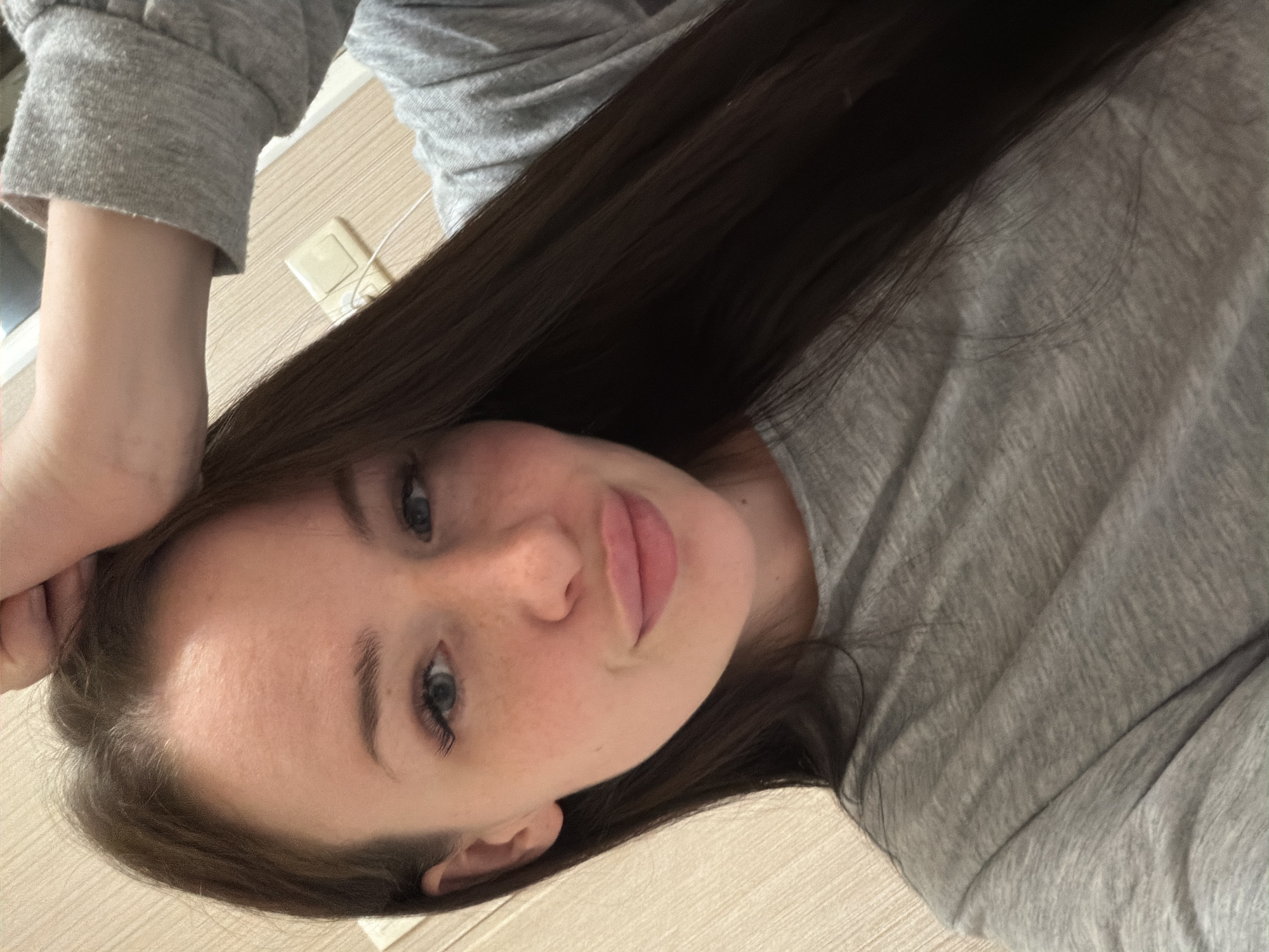 A young woman with long, straight dark brown hair, blue eyes, and a fair complexion with freckles is depicted in a close-up, indoor photograph, identified as taken in the city of Saint Petersburg, Russia. She is wearing a plain, light grey, long-sleeved top. Her lips are full and exhibit a slightly pursed expression, and she looks directly forward with a subtle smile. Her right hand is raised and rests on the upper part of her head. The background consists of a light-colored wall with vertical paneling, and an electrical outlet with a white cable plugged into it is visible behind her right shoulder.