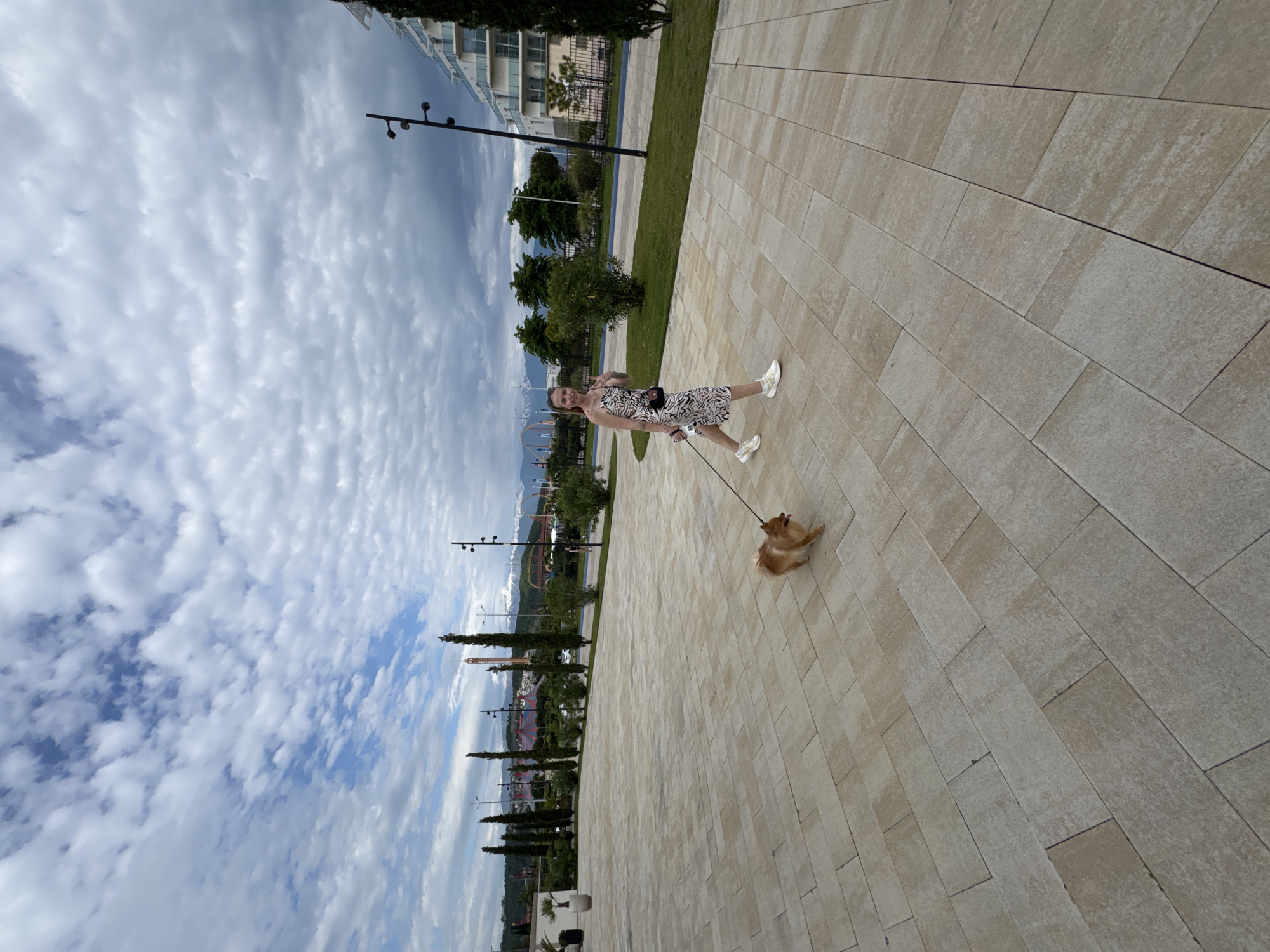 A woman walks a dog on a paved path in Sirius, Russia. The woman, wearing a zebra-print dress and white sneakers, holds a leash connected to a small, light-brown dog. The path is made of large, rectangular light-beige tiles. In the background are trees, buildings, lampposts, and, in the distance, mountains under a cloudy sky.