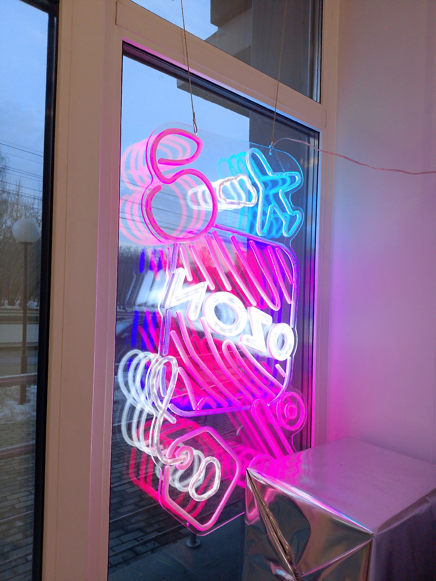 A close-up shot shows a brightly illuminated neon sign hanging in a window in Izhevsk, Russia. The neon sign features a combination of pink, white, and blue lights forming abstract shapes, including what appears to be a stylized "8," an airplane, and the word "Mojo" vertically arranged. The sign is suspended by thin cords.
Outside the window, the blurred urban landscape of Izhevsk is visible. The light suggests the scene is dusk or twilight. The environment appears cold, as indicated by the snow on the ground visible through the window. A modern-style street lamp is also noticeable in the outside scene.
Inside, a silver-covered object resembling a table or box stands to the side, reflecting some of the light from the sign. A white wall is adjacent to the window. The overall mood is modern and potentially commercial, indicating this might be the window display of a business.
