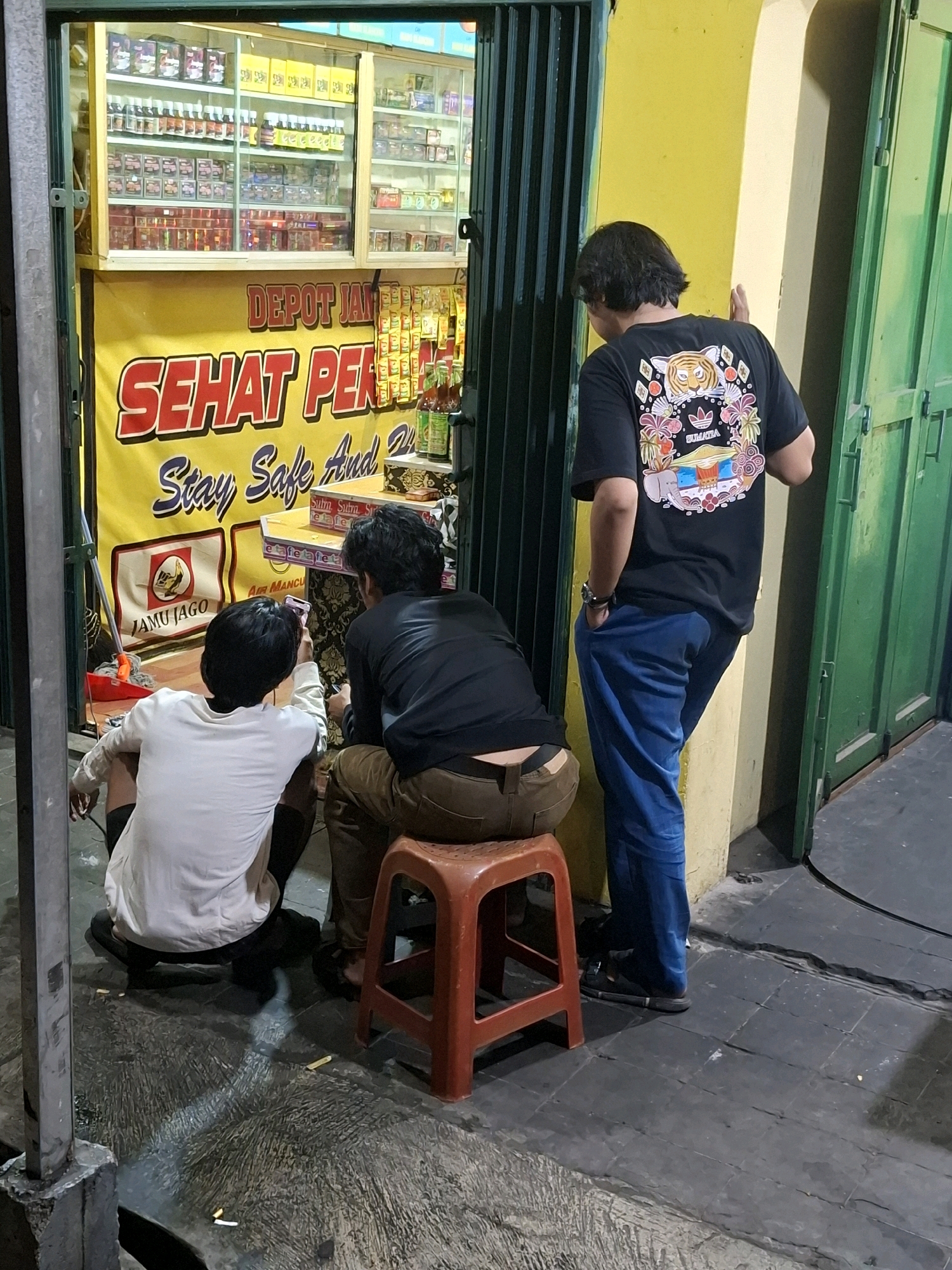 The image depicts an outdoor scene in front of a shop or establishment in Cianjur, Indonesia. In the foreground, two young men are seated on the ground, facing a smartphone. One is wearing a white t-shirt and dark shorts, and the other is wearing a black jacket and brown pants. A third young man, dressed in a black t-shirt and blue pants, stands with his back to the camera, leaning against a yellow wall. The shop behind them has shelves stocked with various products, and a large yellow banner with red and blue text is prominently displayed. The ground appears to be wet and littered with cigarette butts. The overall lighting suggests it is evening or nighttime.