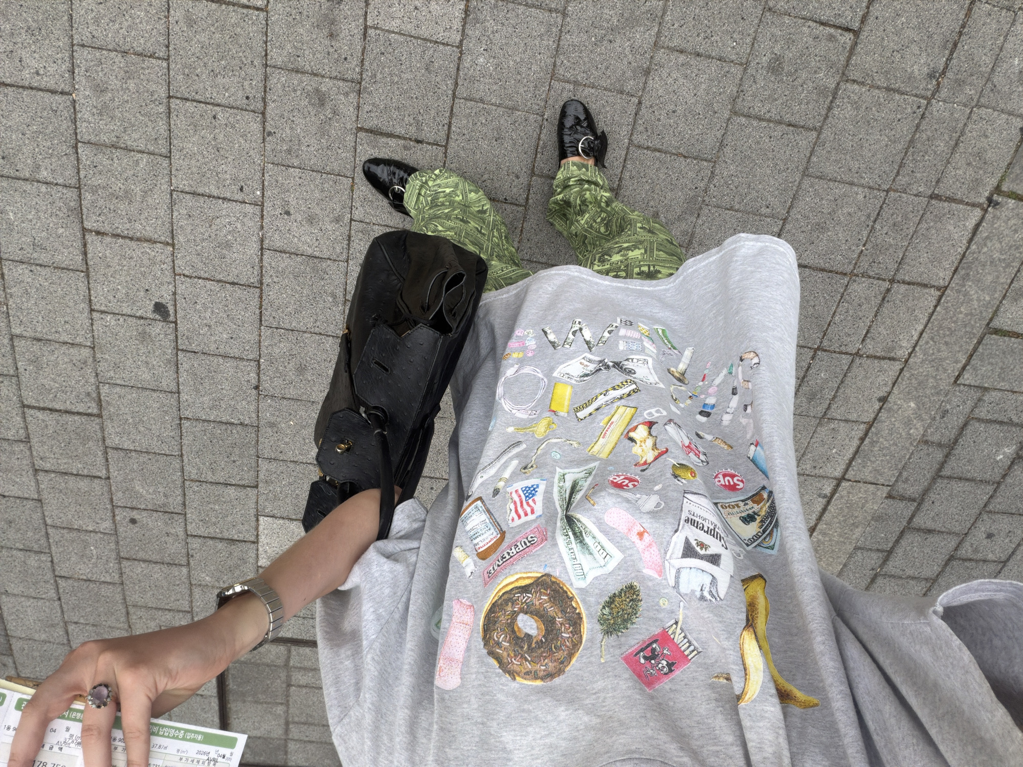 A top-down shot shows a person standing on a gray brick sidewalk. The person wears green pants patterned with money, black shiny shoes, and a gray t-shirt with various images printed on it, including a donut, a banana peel, money, and a pack of cigarettes labeled "Supreme." The person holds a black purse on their left side and a document with Korean text in their left hand. The text includes "리비 납입영수증 (입주자용)" and "2026년 04월." The overall composition suggests a casual, urban scene. The location is likely in the city of Seoul, South Korea based on the Korean text.