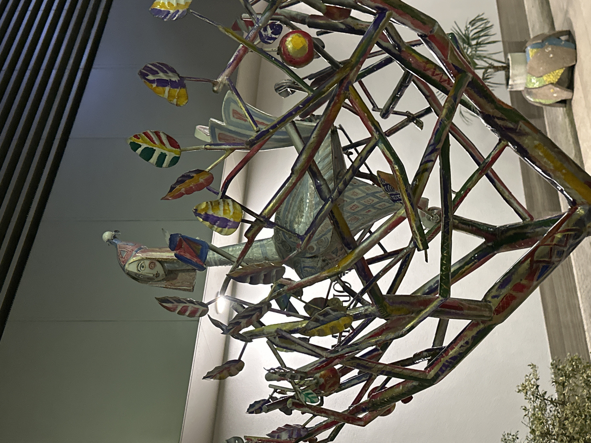 The image depicts a close-up view of a brightly colored, seemingly handcrafted mobile or sculpture. The sculpture features a complex network of painted metal rods and decorative elements, including painted leaves, bird-like figures, and human-like figures. The construction appears somewhat whimsical and folk-art-inspired. In the background, there are glimpses of a building facade. It is not possible to ascertain with certainty from the image if the location is Sochi, Russia.