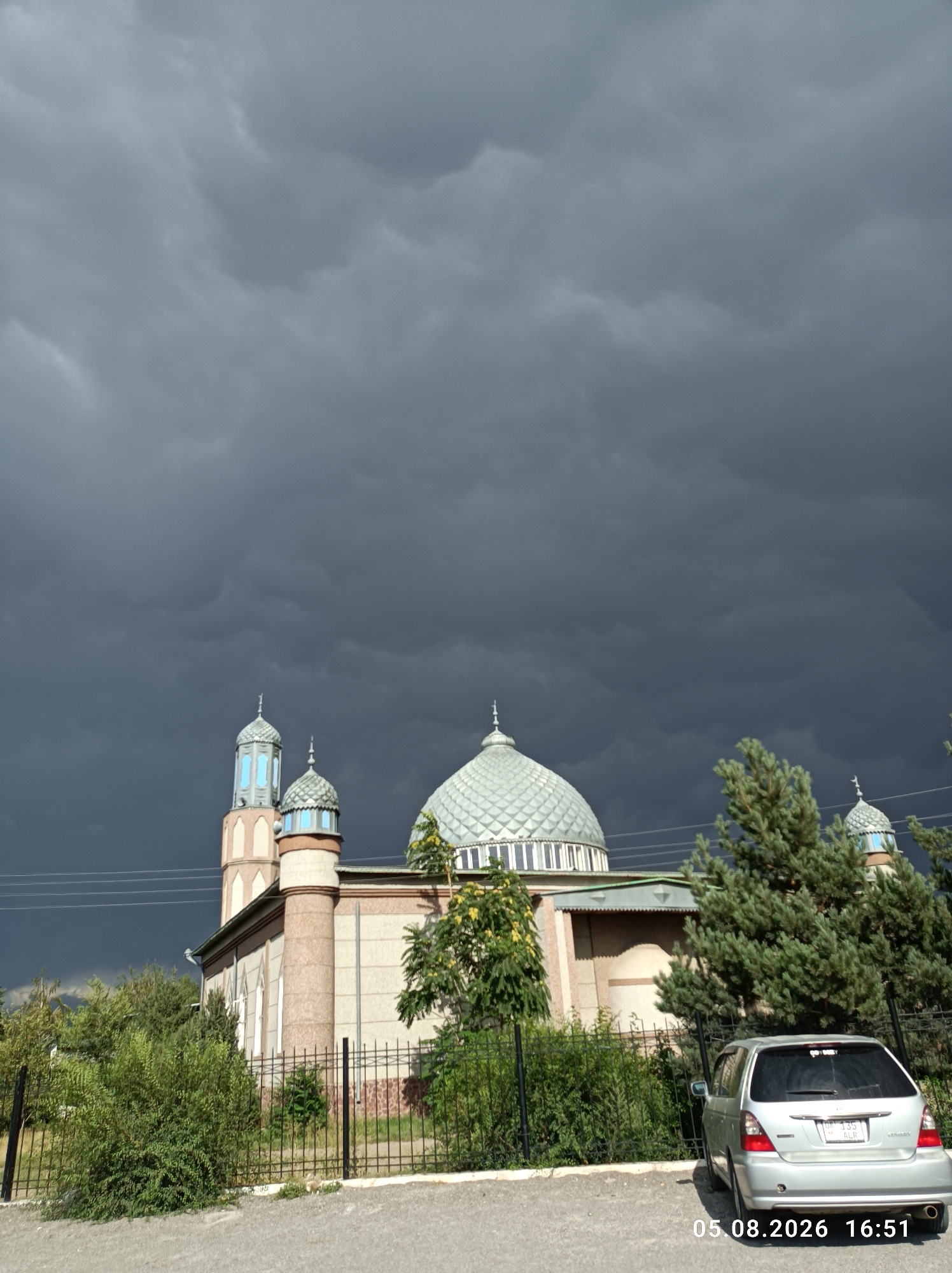 A silver hatchback is parked on a paved surface in front of a mosque. The mosque has a large central dome and two minarets, one of which is visible in the foreground. The sky is dark and cloudy, suggesting an approaching storm. Lush green trees and bushes surround the mosque and are behind a black metal fence. The timestamp "05.08.2026 16:51" is visible in the lower right corner of the image. The scene is likely Chuy, Kyrgyzstan.