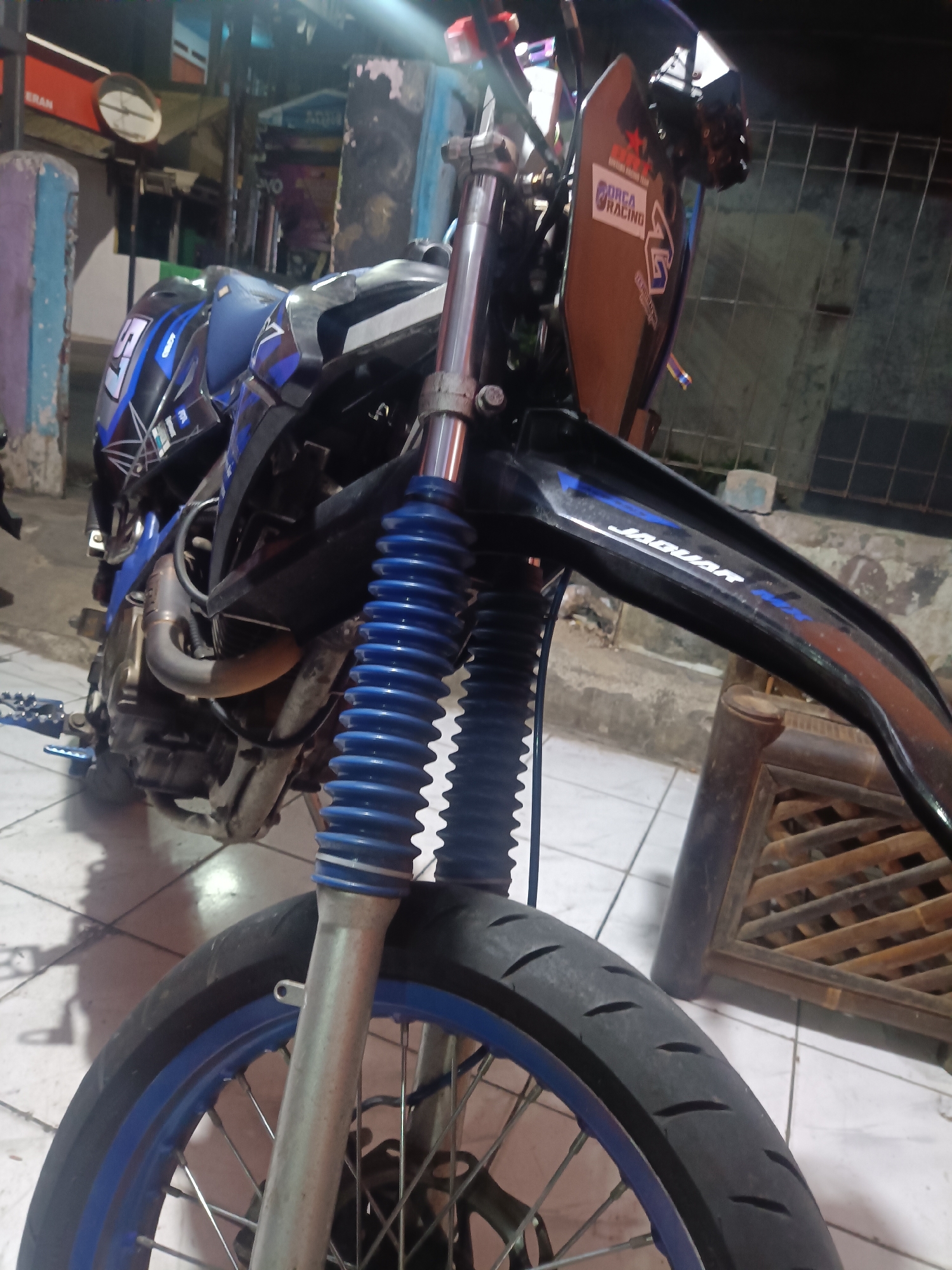 The image shows a close-up, low-angle view of the front of a blue and black dirt bike, parked indoors. The motorcycle has prominent blue front suspension boots. The tire is visible, as is the blue rim and spokes of the front wheel. A black fender extends over the wheel, with a sticker that reads "ORCA RACING". Below the fender, text on the bike's body reads "JAGUAR". The background is dimly lit and appears to be a cluttered indoor space, possibly a garage or workshop, with tiled flooring. In the upper left of the image, a circular clock face with a white background and black numbers is visible on a red background, suggesting it might be part of a shop or establishment. The lighting suggests it is nighttime or the indoor space is poorly lit. No people are visible in the frame.