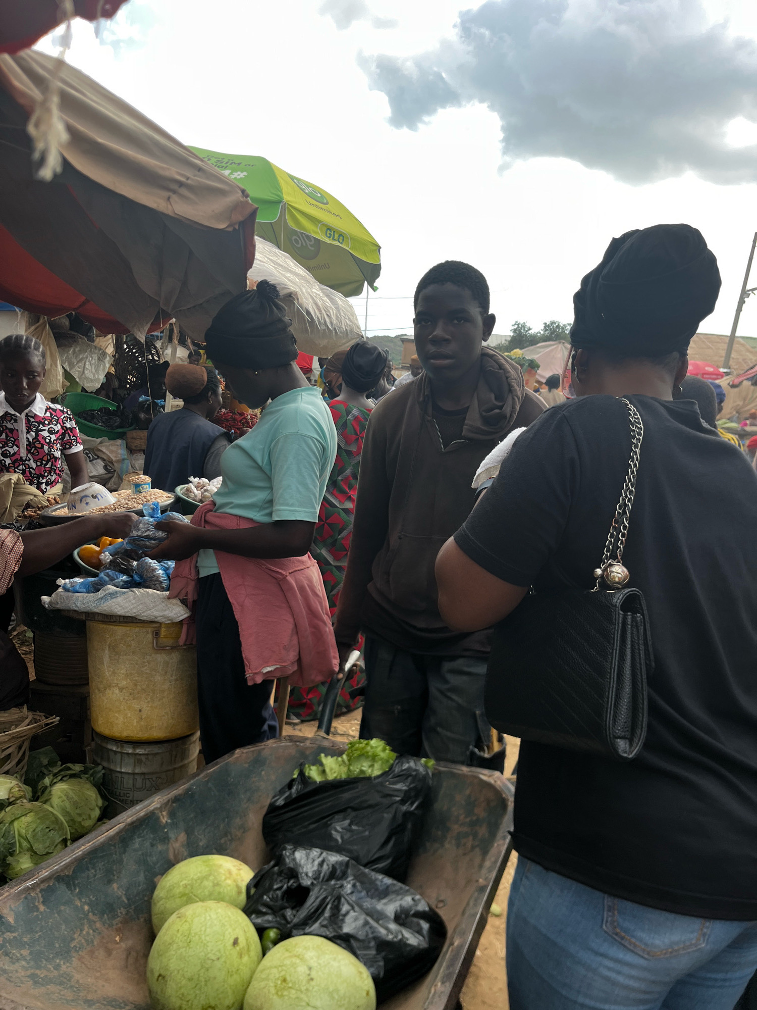 A medium shot shows a bustling outdoor market scene in Gura Top, Nigeria, under a cloudy sky. In the foreground, a wheelbarrow is filled with produce, including several large green watermelons and a bag of leafy green vegetables. To the left of the wheelbarrow, cabbages are piled up. Behind the wheelbarrow, several people are gathered around market stalls.
A young man with dark skin and short hair, wearing a brown hooded jacket, stands looking forward. To his right, a woman wearing a black headwrap and a black t-shirt is seen from behind, with a black handbag with a silver chain strap slung over her shoulder. She is wearing blue jeans.
In the middle ground, a woman in a light blue t-shirt and a pink shawl draped around her shoulders is holding plastic bags containing items, possibly food. She is standing behind a stall with oranges and other items in bowls. Another woman with a black headwrap and a patterned top stands to her left, looking towards the stall. Other market-goers and stalls are visible in the background. The market appears to be covered by various tarpaulins and umbrellas, including a green umbrella with "GLO" branding. The ground is dirt.