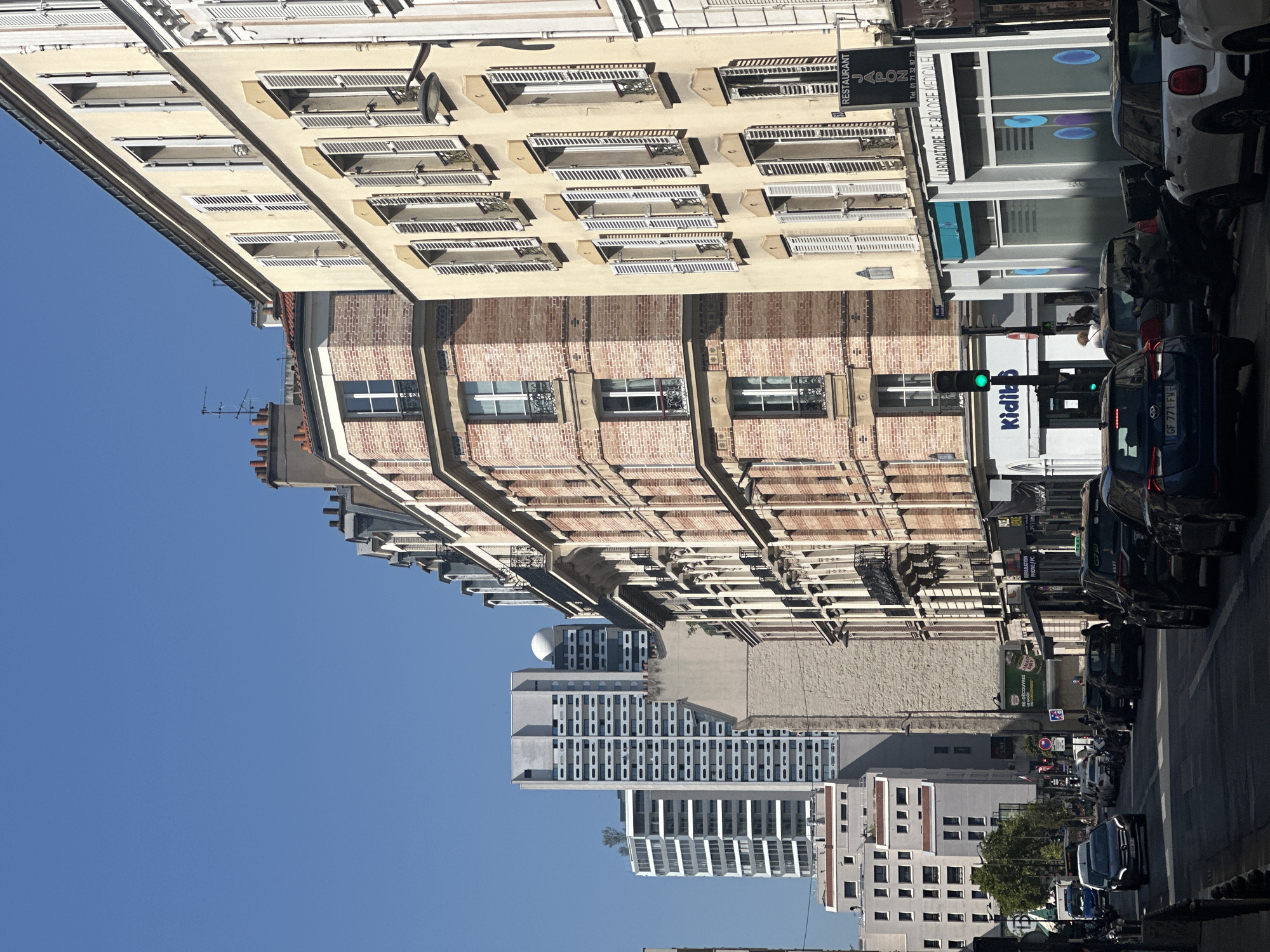 The image depicts a street scene in Paris, France, likely on a sunny day, given the clear blue sky and the shadows cast by the buildings. The street is lined with a mix of architectural styles, ranging from traditional Parisian Haussmannian buildings with brick facades and mansard roofs to more modern, taller structures. The foreground shows a street with several cars, including a dark blue Mercedes-Benz, a dark grey SUV, and a white Mini Cooper, suggesting moderate traffic. Pedestrians are visible on the sidewalks, and some are entering or exiting buildings.
The buildings on the right side of the street are predominantly light yellow with cream-colored shutters, many of which are open, revealing glimpses of windows. Some of these buildings have businesses on the ground floor, indicated by signage such as "RESTAURANT JAPON" and "LABORATOIRE DE BIOLOGIE MÉDICALE". A traffic light is visible and appears to be green for the direction of the cars in the foreground.
Further down the street, taller, more contemporary buildings are visible. One prominent building is a tall, light grey structure with numerous windows arranged in a grid pattern. Another building to its left is a white, glass-fronted skyscraper. The presence of a mix of older and newer architecture suggests an area undergoing development or that is a transition zone between historical and modern districts within the city. The overall impression is one of an active urban environment.