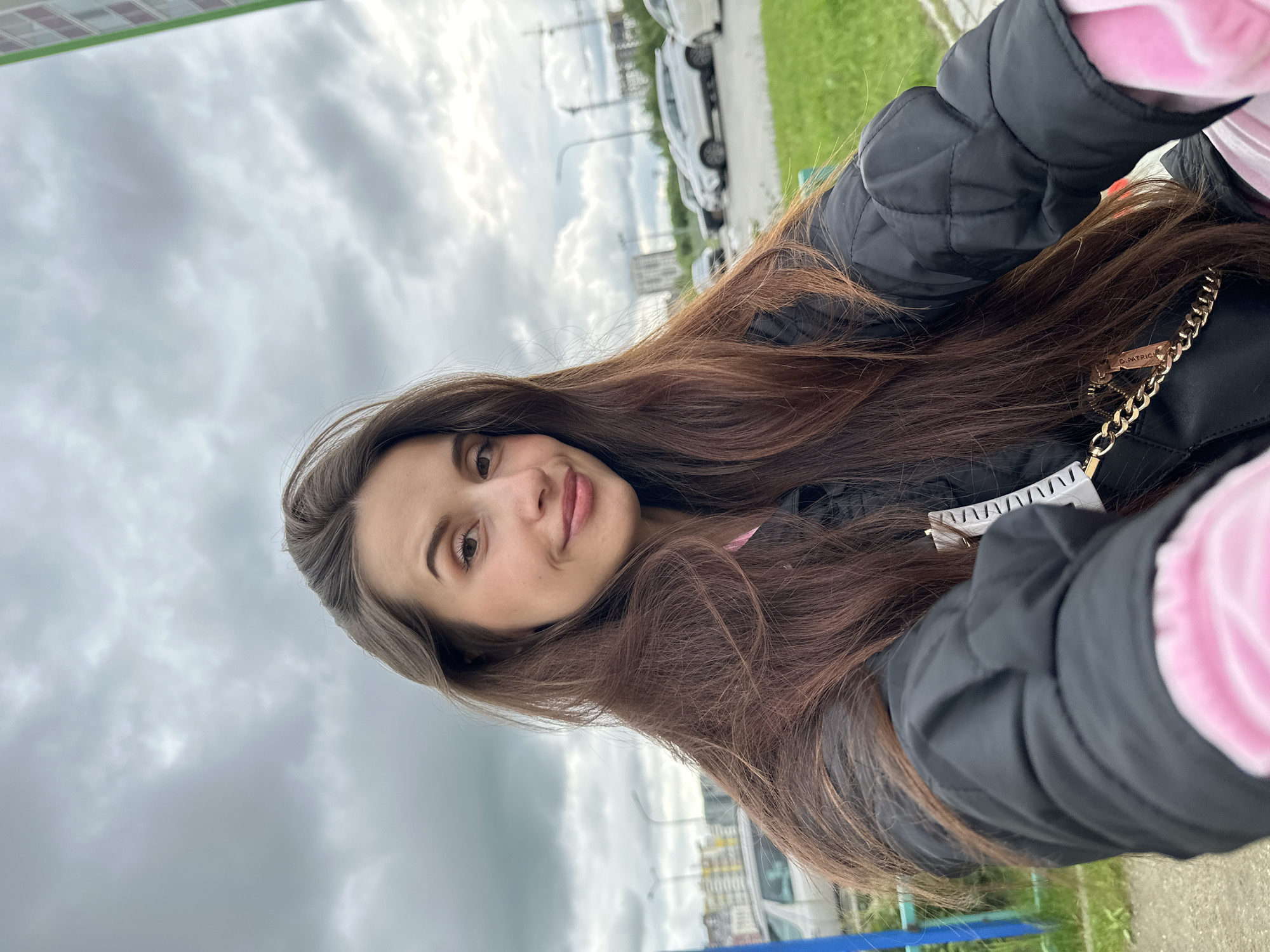 A young woman with long, dark brown hair, wearing a black quilted jacket over a pink top, takes a selfie outdoors. Her hair is partially obscuring her face, which is tilted slightly towards the camera, and she is smiling faintly. She has noticeable makeup on her eyes and lips. She's wearing a gold chain purse. The background shows a grey, cloudy sky, some buildings under construction in the distance, parked cars, a patch of green grass, and a blue railing. This appears to be a typical urban environment.