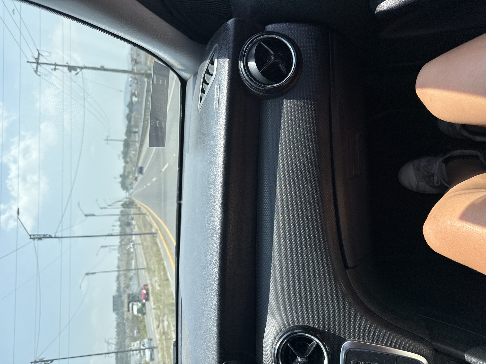 The image shows an interior view from the passenger seat of a vehicle, possibly a car, traveling along a highway near San Francisco Totimehuacán, Mexico. Outside the window are power lines and light posts lining the road, and there is evidence of other vehicles. Inside, a textured grey dashboard is prominent, with air vents visible. A portion of a person's legs, clad in light-colored clothing, and feet in grey shoes are visible on the right side of the image.