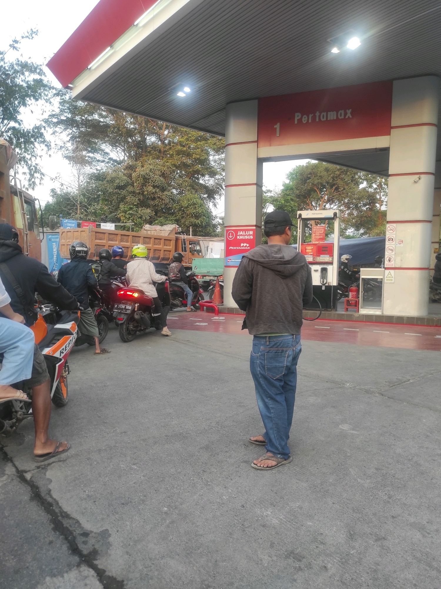 This is a full-shot photograph of a Pertamina gas station in Padalarang, Indonesia, captured outdoors during daytime. The scene depicts a queue of motorcycles and their riders at the fuel pumps, with a large truck visible in the background. The foreground features a man wearing a grey hooded jacket, blue jeans, and sandals, standing with his back to the camera, facing the gas station. To his left, another individual, partially visible, is wearing blue pants and sandals, sitting on a motorcycle. Several other individuals on motorcycles are also present, some wearing helmets in yellow, blue, and black. The gas station itself has red and white branding with the Pertamax logo prominent. The ground is paved asphalt, with some wet patches visible. Trees and foliage surround the gas station, indicating a somewhat rural or semi-urban setting. The overall impression is of a busy, functional gas station during a period of refueling activity.