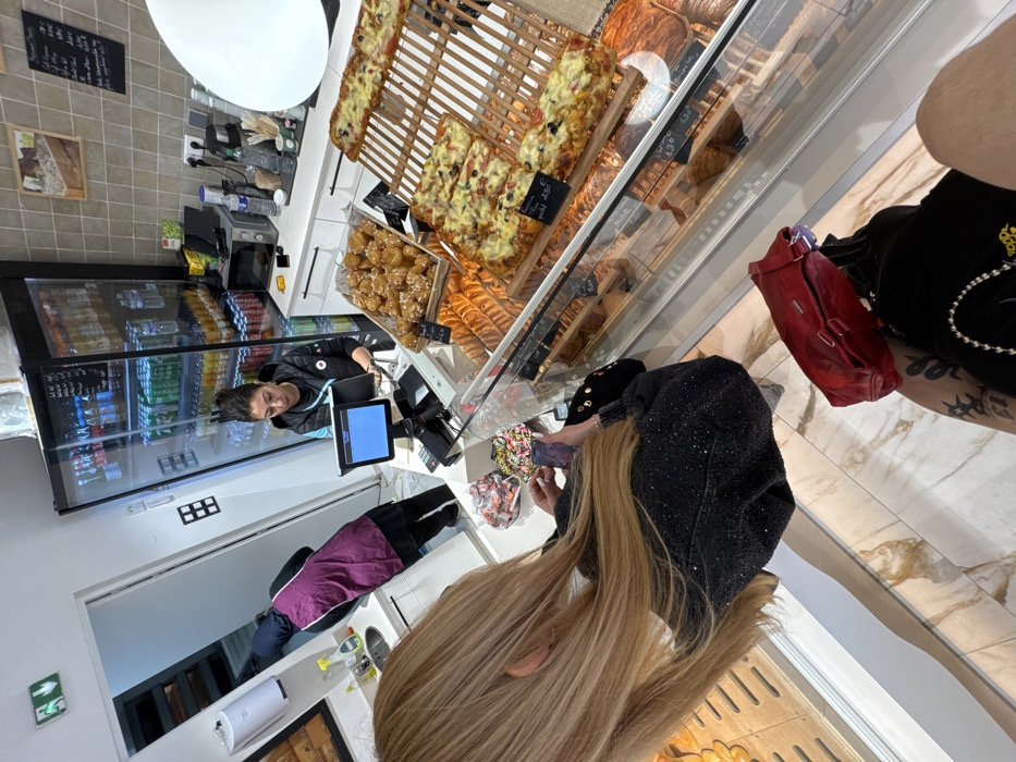This image depicts the interior of a commercial establishment, likely a bakery or snack shop, located in Asnières-sur-Seine, France. In the immediate foreground, partially visible from behind, a person with long blonde hair wears a dark, possibly sequined jacket. This individual holds a smartphone in their left hand, oriented as if capturing an image or video. A red handbag rests beside their right leg, which displays a tattoo. Behind a glass display counter, a female employee with dark hair pulled back, wearing a black top with a logo, stands at a point-of-sale terminal, which features a blue screen. To her right, a tall, glass-fronted refrigerator is stocked with various bottled and canned beverages. The counter behind her is white and includes a microwave oven and beverage supplies. The display counter showcases a large basket of golden-brown, glazed pastries and multiple rectangular flatbreads, resembling pizza slices, arranged on wooden stands with price labels, including one indicating "2,50 €". Below the visible food, further pastries are displayed within the glass case. In the background, through an open doorway, a second employee is partially visible, wearing a dark garment and a purple apron, positioned near a sink area. The establishment features light-colored, square wall tiles and a patterned marble floor. An emergency exit sign is affixed above the doorway.