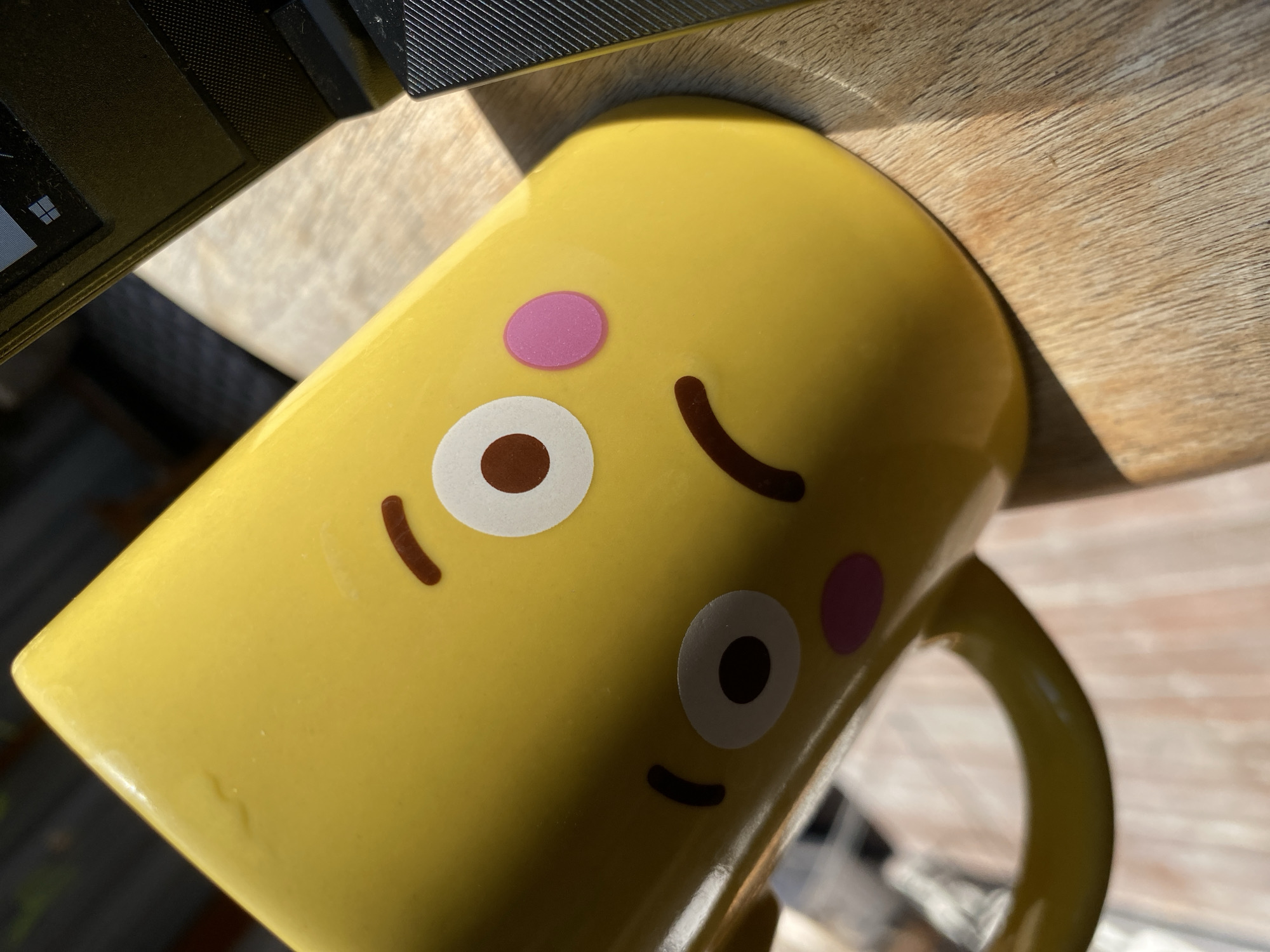 A yellow mug with a cartoon face is placed on a wooden surface. The mug is tilted, showing its front with two eyes, a mouth, and two pink circles on its cheeks. Part of a black laptop is visible on the right side of the image. The scene appears to be outdoors, with natural light casting shadows on the mug and the wooden surface. The background is out of focus but suggests an outdoor environment with some vertical structures. There are no people or animals present in the image. The overall setting appears to be casual, possibly a desk or table outdoors. The image was taken at a slightly elevated angle, looking down at the mug.