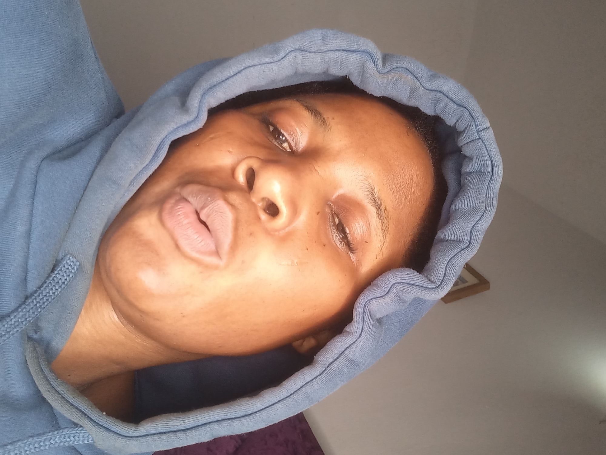 A young woman wearing a blue hoodie poses for a close-up selfie. Her eyes are partially closed, and her lips are pursed. The background is a plain wall with a framed picture on it. The lighting suggests an indoor setting, possibly a home. No specific location in Jos, Nigeria is discernible from the image itself.