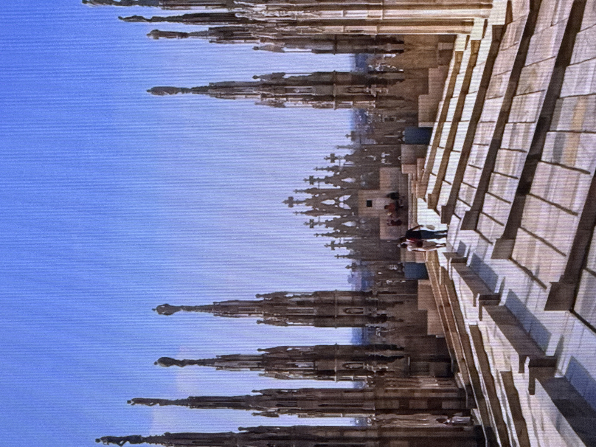 This is an image of the Milan Cathedral's rooftop in Italy. The rooftop features a complex architectural design with numerous spires and statues. In the foreground, the surface is paved with stone slabs, forming a tiered walkway. Two individuals are walking away from the camera on this walkway, while other people are visible in the background, seated and standing near the architectural structures. The sky is a clear, bright blue. The image does not depict any part of Dubai, United Arab Emirates.
