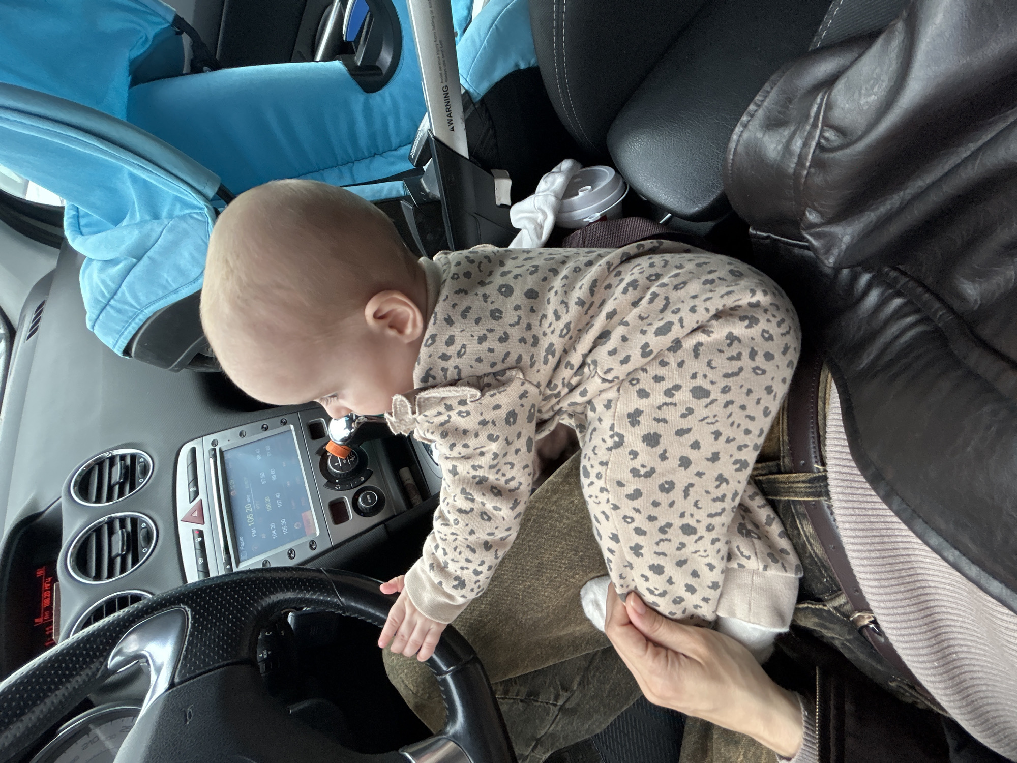 A baby in a leopard-print onesie is reaching for the steering wheel of a car. The baby is being held by an adult whose hand is visible. The car's dashboard and radio are visible, displaying "FM 106.20 MHz" and a radio frequency. Part of a child's car seat can be seen. There's also a sign with "AWARNING" on it. The location is likely inside a car, and based on the Cyrillic letters on the radio, possibly in a Russian-speaking country, potentially Moscow, Russia.
