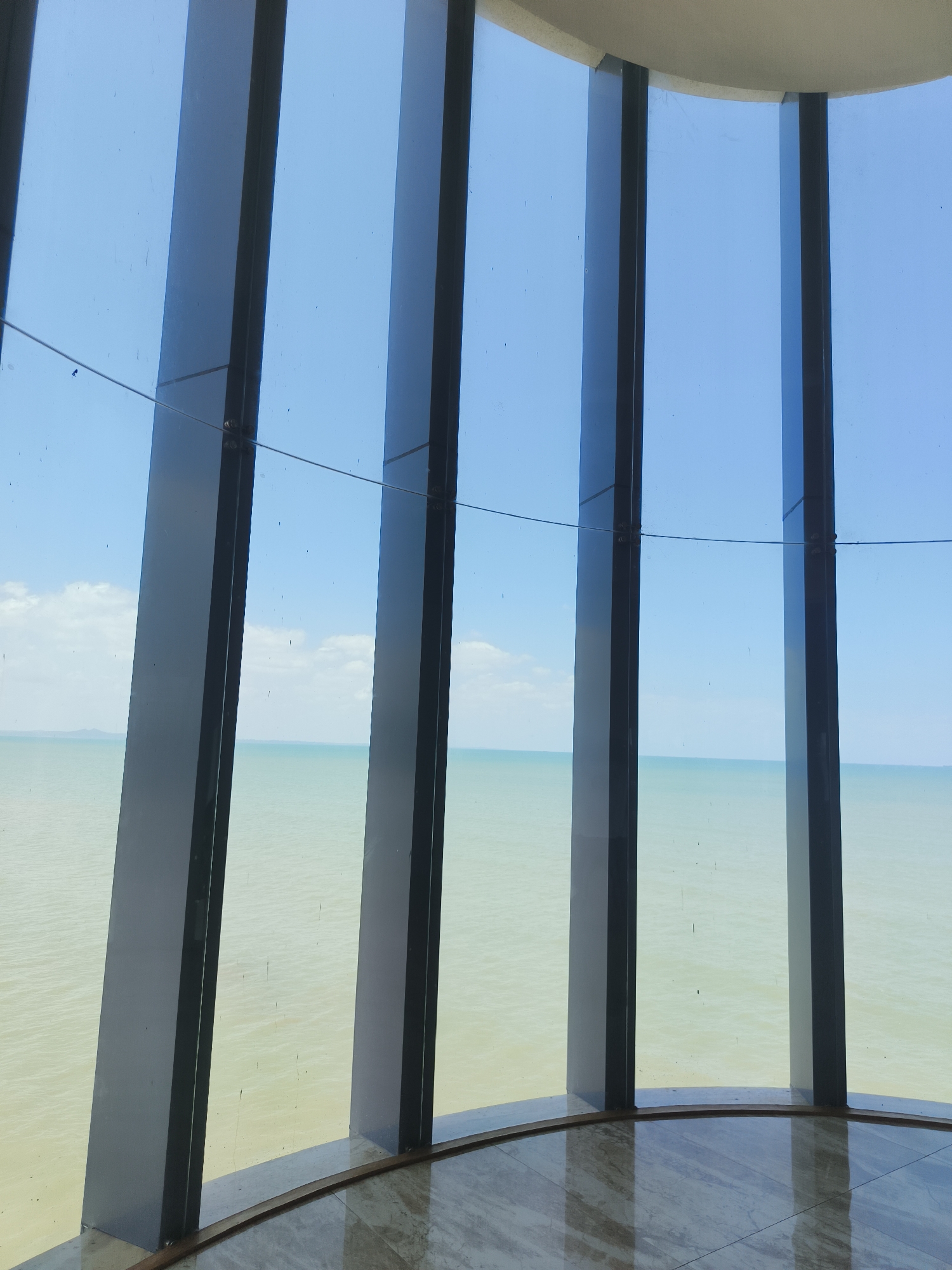 The image captures a view of a calm, pale blue sea and sky through a large, curved window with dark vertical supports. The window frames are dark gray and angled, suggesting a modern architectural design. The floor appears to be polished stone or marble, reflecting the light and the window supports. In the distance, a faint line of land is visible on the horizon. The sky is clear with a few wispy clouds. There are no people or objects in the immediate foreground or on the sea. The overall impression is one of serenity and expansive natural beauty viewed from an indoor vantage point.