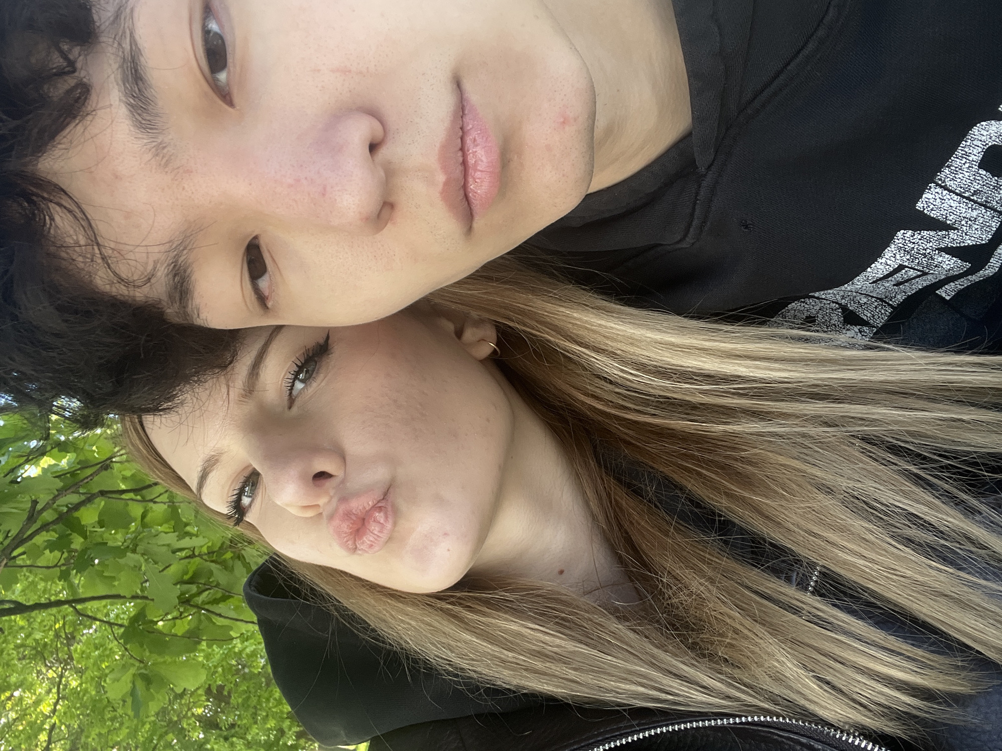 The image shows two individuals, presumably a couple, in an outdoor setting in the city of Unknown, Unknown.
The person on top has dark hair and is wearing a black hooded sweatshirt with white lettering. Their face is close to the camera, showing facial features.
The person below has blonde hair and is also close to the camera, also showing facial features, including pursed lips as if giving a kiss. They are wearing a dark jacket with a metal zipper detail.
Green foliage and trees are visible in the background, suggesting a park or outdoor area. The overall composition appears to be a selfie.
