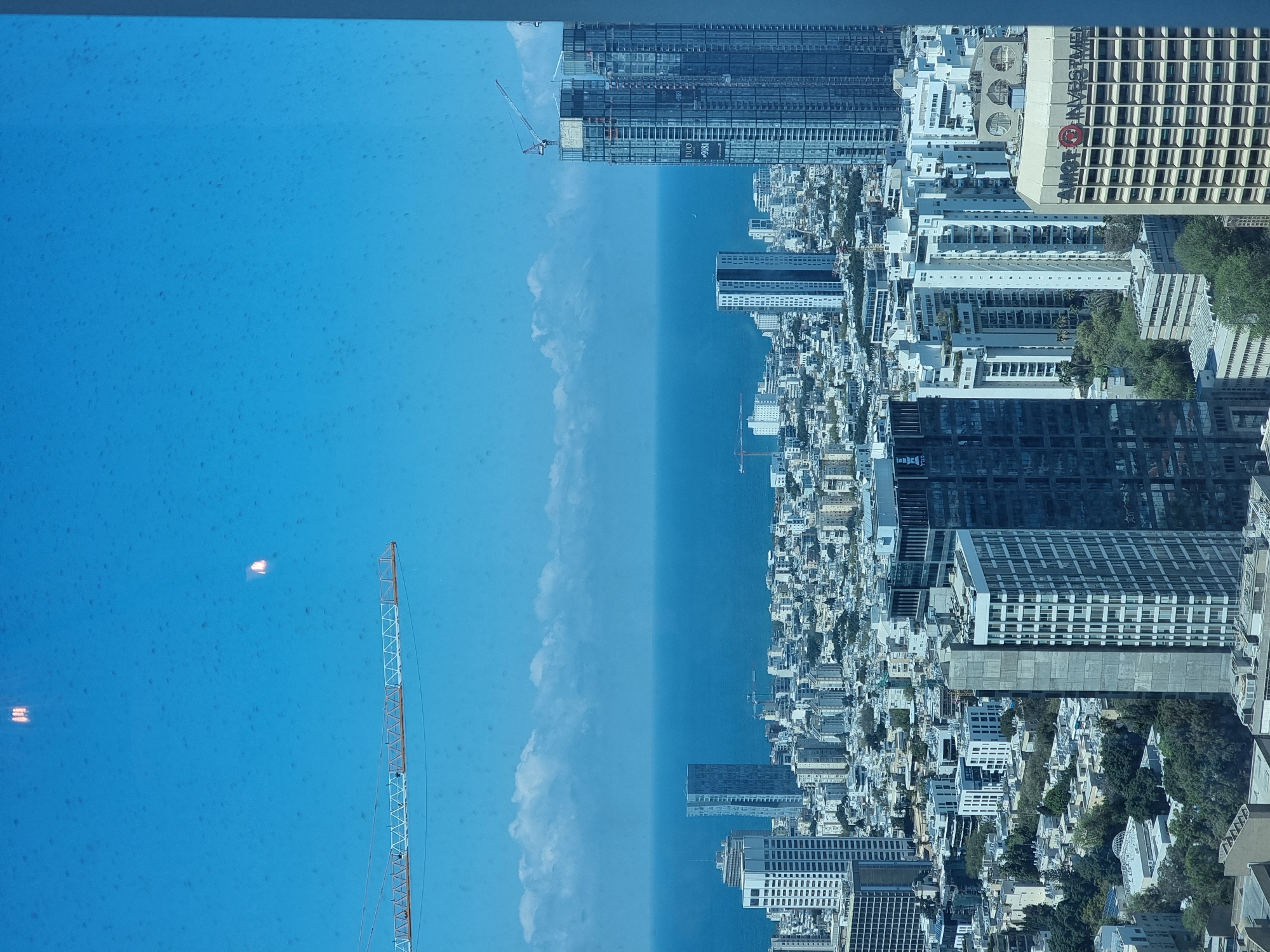 The image presents a high-angle, slightly blurry view of Tel Aviv, Israel, likely captured through a window. The left portion of the frame is dominated by a bright blue expanse representing the sky and possibly the Mediterranean Sea, interspersed with subtle cloud formations. Two construction cranes are visible in this section. The right side of the image reveals the urban landscape of Tel Aviv, characterized by a dense arrangement of buildings ranging from residential structures to high-rise office towers. Prominent buildings are "Amot Investments" building. The blurred nature suggests the image was taken from an elevated position, potentially inside a skyscraper or from high above the city.