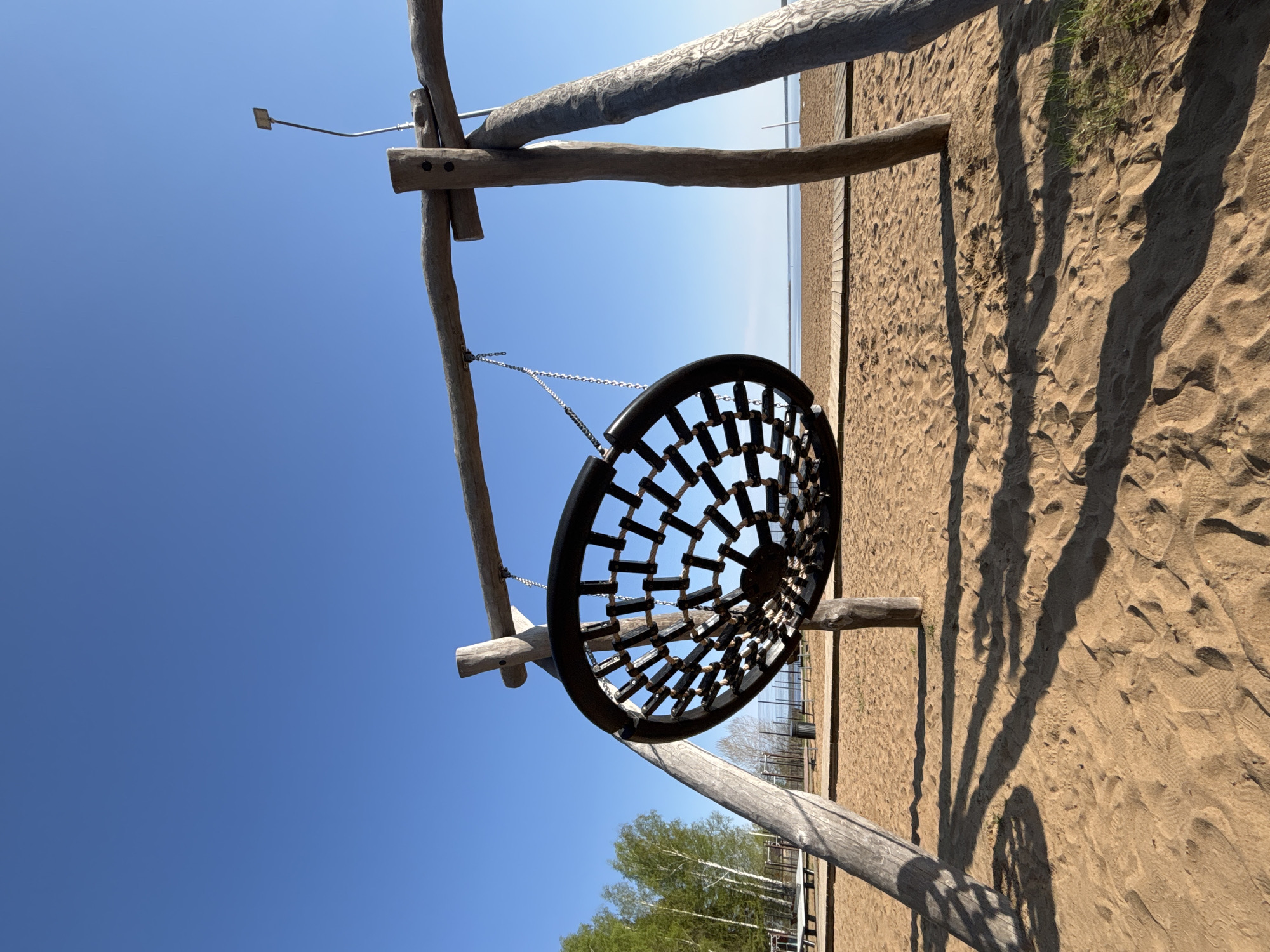The image depicts a playground structure on a sandy surface under a clear blue sky. The structure is made of wooden logs and includes a round swing suspended by chains. The swing has a dark-colored frame with a grid-like pattern in the center. The sandy surface shows various footprints and shadows. The background includes vegetation, possibly trees, and a distant horizon line, suggesting a body of water. The image was taken in the city of Chaikovsky, Russia.