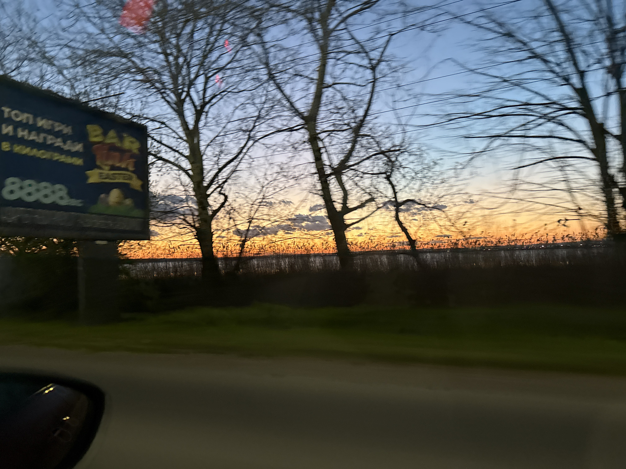 The image shows a view from inside a vehicle, likely a car, at dusk or dawn. The sky is a blend of orange and blue hues, indicating either sunrise or sunset. Silhouetted trees stand against the sky, their bare branches creating a stark contrast. In the foreground to the left is a large billboard advertising a BAR, with some lettering in Cyrillic script. The sign also has the word "EASTER" in English. Below the billboard, there is a stretch of green grass. In the distance, beyond the trees, there is what appears to be a body of water bordered by reeds or tall grasses. The road is in the foreground and part of a car's side mirror is visible in the bottom left corner. While a precise location within Burgas, Bulgaria, is difficult to pinpoint without additional landmarks, the scene suggests a roadside view on the outskirts of the city, possibly near a lake or body of water. The image captures a moment of transit at twilight, marked by the billboard's advertisement and the natural beauty of the sky and surrounding landscape.