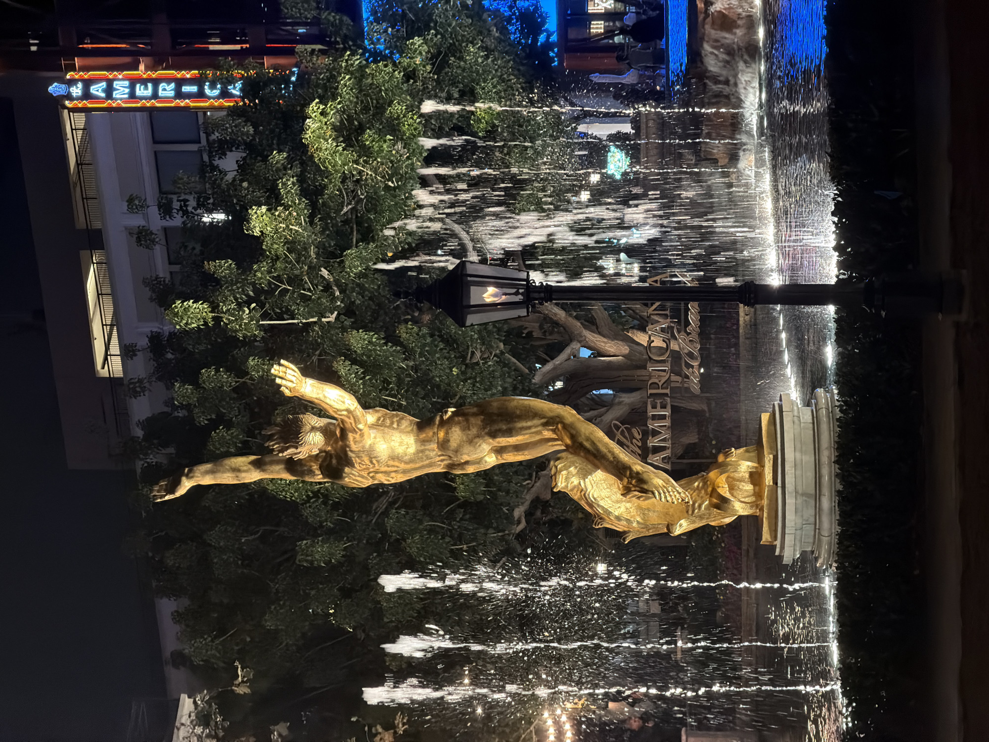 The image shows a nighttime view of the fountain at The Americana at Brand in Glendale, California, United States. The fountain is centered in the frame with a golden statue of a nude male figure diving into the water. Water sprays from the fountain in multiple streams, catching the light. Behind the statue are trees and a lamppost with a flickering flame. The sign for "The Americana at Brand" is partially visible, as is a building with lit windows in the background. The sky is dark blue.