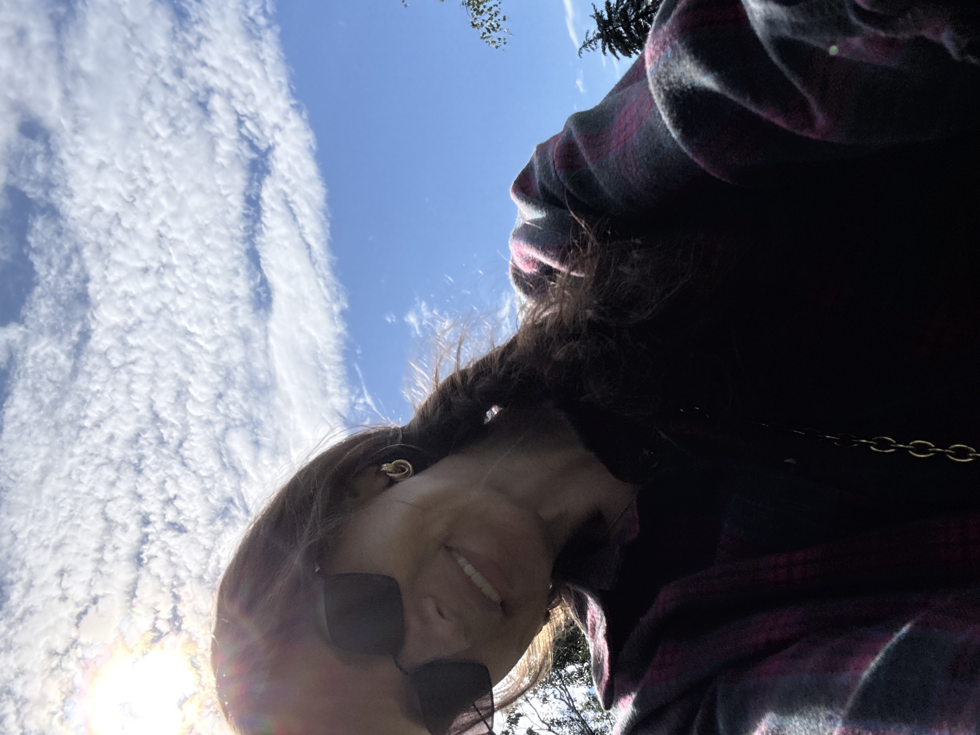 The image is a selfie taken outdoors, looking up at the sky. The sun is bright, casting lens flare and illuminating wispy clouds against a clear blue sky. A person, only partially visible, is wearing a dark plaid jacket with pink and gray accents and dark sunglasses. They have dark hair and a gold earring. Their mouth is open in a smile, suggesting a happy or cheerful emotion. The overall impression is of a bright, sunny day. The scene is likely in a park or other outdoor area, with some foliage visible in the background. The time of day appears to be daytime, given the strong sunlight and blue sky. There is no visible text in the image.