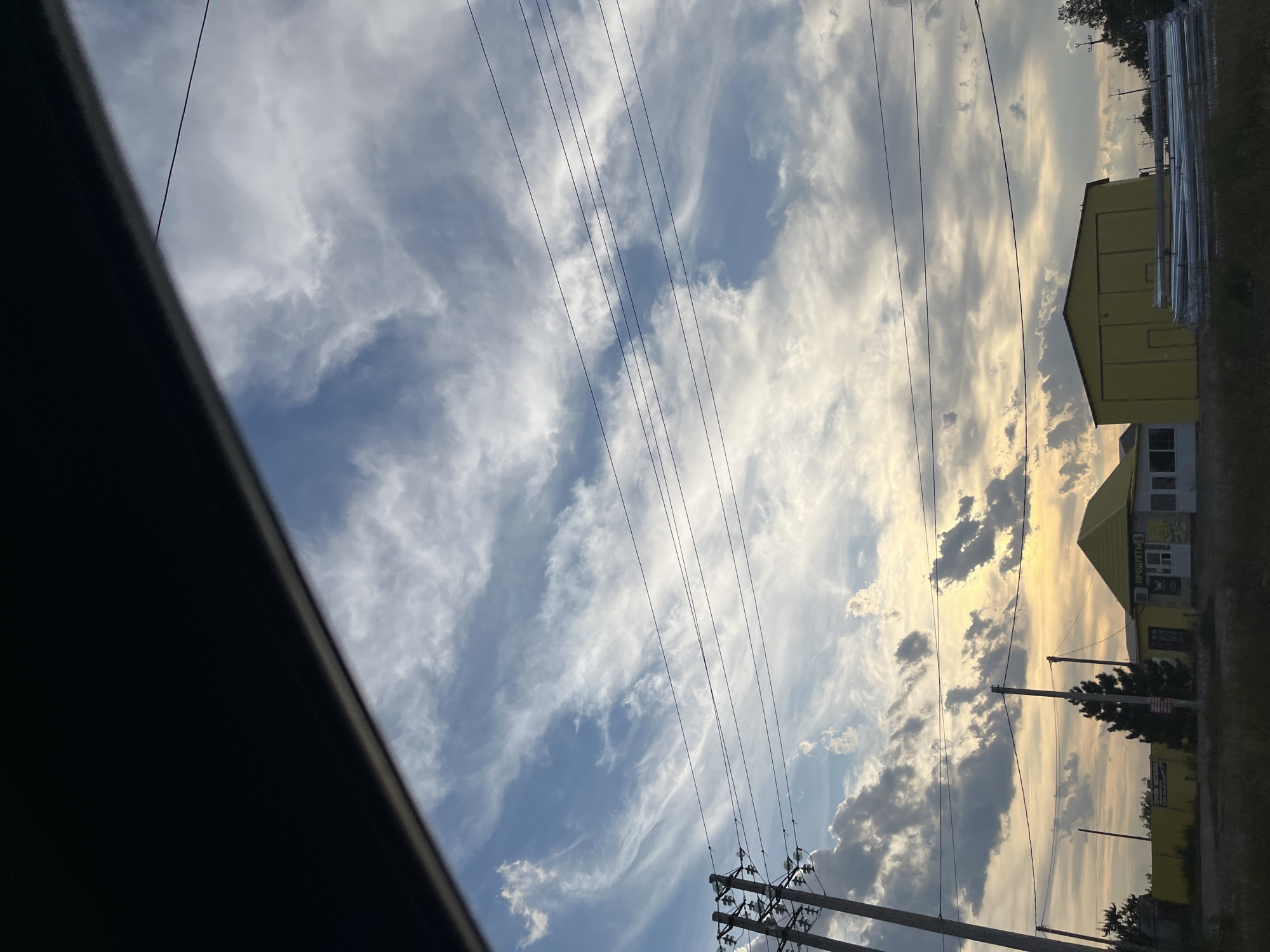 The image depicts a view through a dark, angled frame, likely a car window, looking out at a cloudy sky. The sky is a mix of blue and white clouds, with streaks of cirrus clouds creating a wispy effect. In the distance, there are several buildings, a fence, and utility poles with overhead wires. The buildings appear to be industrial or commercial, with one building featuring a yellow roof and the Cyrillic text "ПРОДУКТЫ" (PRODUKTY, meaning "products") above a storefront. Another building to the right is also yellow and appears to be a warehouse or industrial shed, with stacks of long, cylindrical objects, possibly pipes, stored in front of it. A solitary evergreen tree stands between the buildings. The lighting suggests it is either sunrise or sunset, with a warm glow to the sky. The scene appears to be set in an outdoor, possibly rural or suburban, area. There are no people visible in the image. The location is identified as the city of Shumeika, Russia.