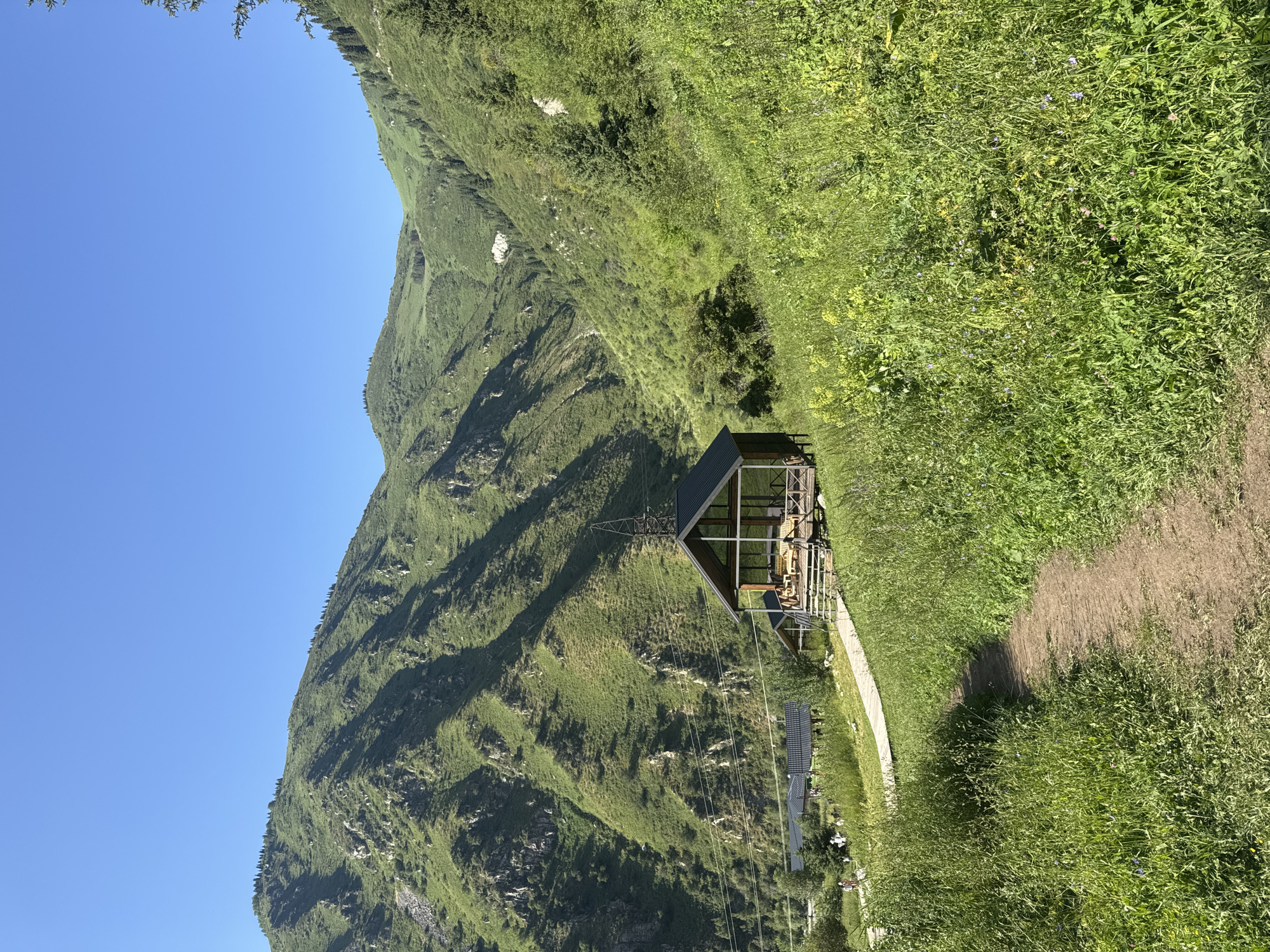 A dirt path winds through lush green grass and wildflowers in the foreground, leading towards a wooden gazebo with a dark roof. The gazebo is situated on a raised wooden platform and appears to be open-sided, with visible wooden benches and tables inside. In the background, several other low-rise buildings with dark roofs are nestled amongst trees. Further beyond these structures, steep, verdant mountains rise dramatically under a clear blue sky. The mountains are covered in dense green vegetation and show signs of rocky outcrops and ravines. The scene suggests a natural park or recreational area in a mountainous region. Based on the landscape, this location is likely in the Ile-Alatau National Park, just outside Almaty, Kazakhstan, a popular area for hiking and outdoor activities with its striking mountain scenery and well-maintained trails.