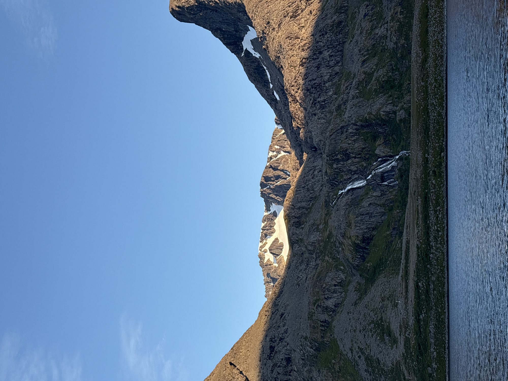 The image depicts a scenic view of the coast in Arnøyhamn, Norway, with dramatic mountains meeting a calm body of water. The sky is clear and blue with a few wispy clouds on the left. The mountains are rugged and rocky, with patches of snow visible on the higher peaks. A waterfall cascades down the mountainside in the middle ground. The foreground shows a body of water, likely the sea, with gentle ripples on its surface. The lighting suggests it is either early morning or late afternoon, with the sun casting long shadows across the mountains. There are no people or man-made structures immediately visible, emphasizing the natural beauty of the landscape.