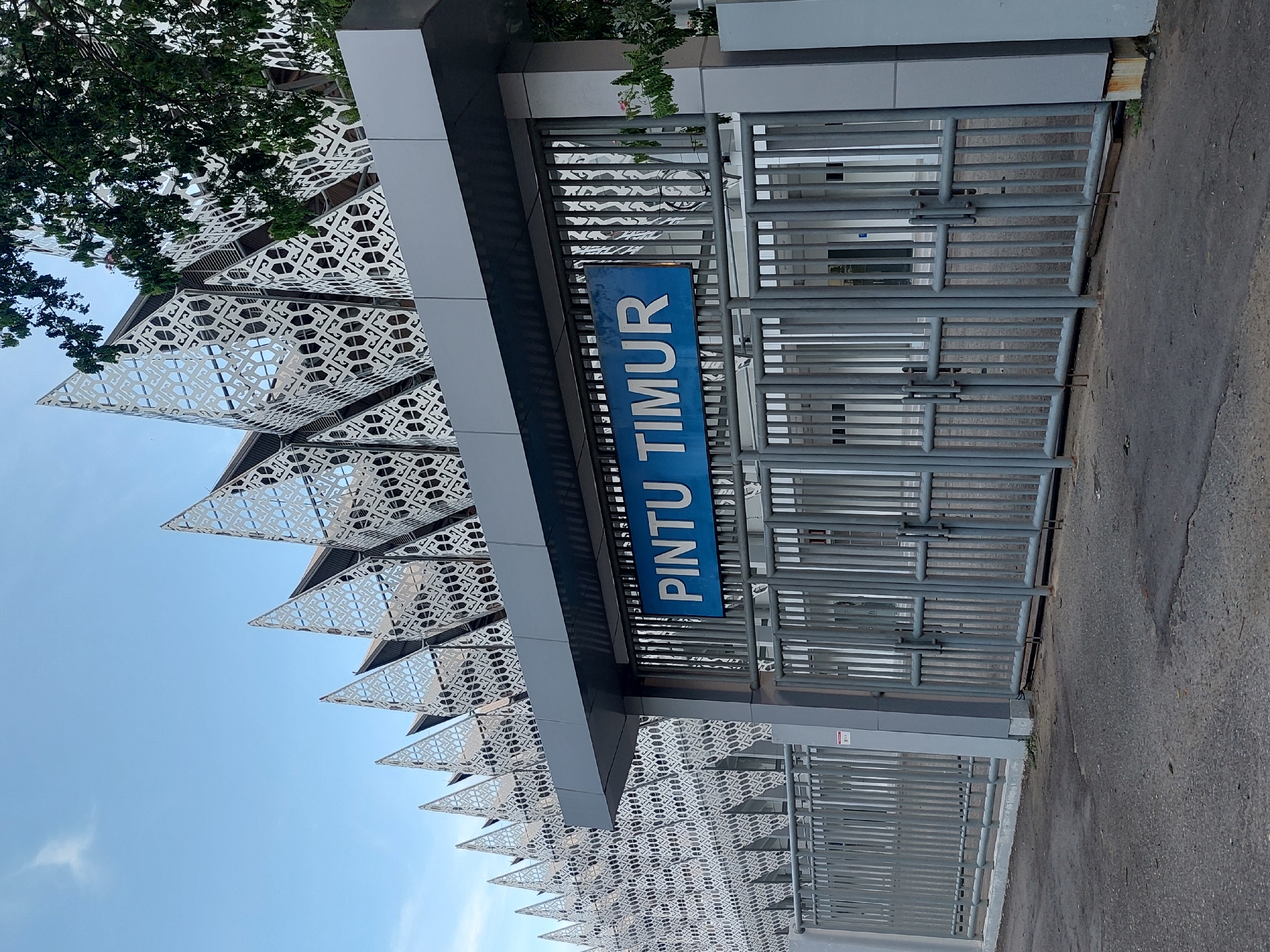 This is an exterior, ground-level, medium shot of the East Gate (Pintu Timur) of a modern building, possibly a stadium or event venue, located in Banda Aceh, Indonesia. The gate is a series of tall, gray metal bars with multiple latches and handles, suggesting it's a secure entrance. Above the gate, a blue sign with white lettering reads "PINTU TIMUR." The building's facade features a distinctive, angular roofline with triangular, perforated metal panels that have an intricate, repeating geometric pattern. These panels extend across the top of the structure, creating a dynamic and decorative effect against the clear, pale blue sky. To the right, a lush green tree with dark leaves partially obscures the building's upper portion. The ground in front of the gate is paved with asphalt, showing some wear and minor debris. There are no people visible in the image, and the scene appears quiet and deserted, as if captured during off-hours or before an event.