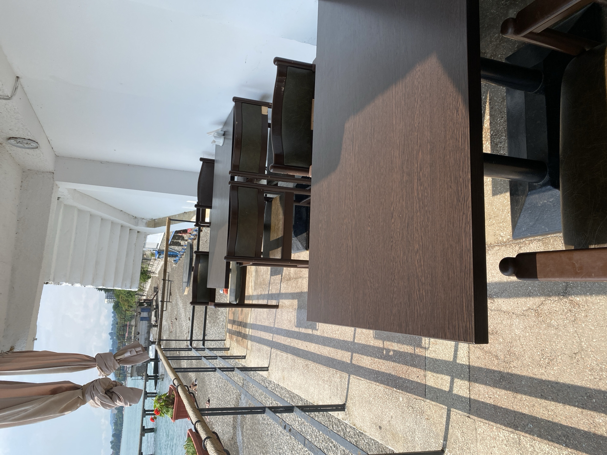 This image depicts an outdoor seating area of a restaurant or cafe, likely in Chabkwara, Abkhazia, overlooking a beach and the sea. In the foreground, a dark brown wooden table is visible, casting a shadow on the textured concrete floor. To the left, a black metal railing separates the seating area from the beach. Beyond the railing, a dark gravel beach stretches to the water, with a pier extending into the sea. Several potted plants with red flowers are placed along the railing.
The seating area itself contains multiple dark brown wooden tables and chairs with green upholstered seats and backs. Sunlight casts long, distinct shadows of the railing and possibly overhead structures across the patio. In the background, a white building with white beams and a corrugated roof is visible, suggesting the establishment's architecture. The sea is calm and a light blue, with a distant shoreline featuring green trees and buildings. The sky is overcast with some visible clouds. No people are clearly identifiable as subjects, although there may be indistinct figures on the beach in the distance. The overall impression is one of a quiet, seaside dining establishment on a sunny day.