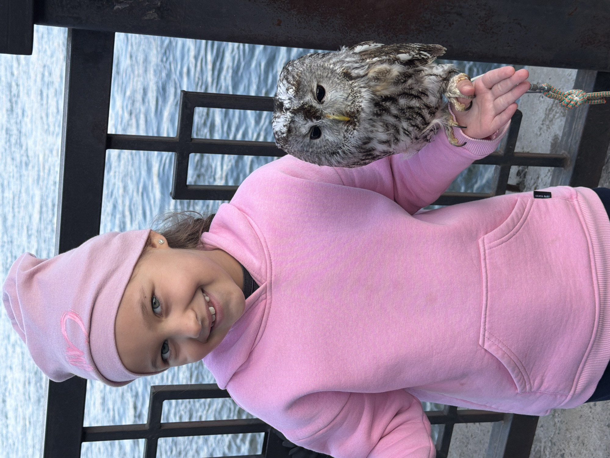A young girl in a pink hoodie and matching hat is holding an owl. The girl is smiling at the camera, and the owl is perched on her left hand. The background shows a railing and water, suggesting they are near a river. Based on the surrounding environment, this scene appears to be captured in Krasnoyarsk, Russia.