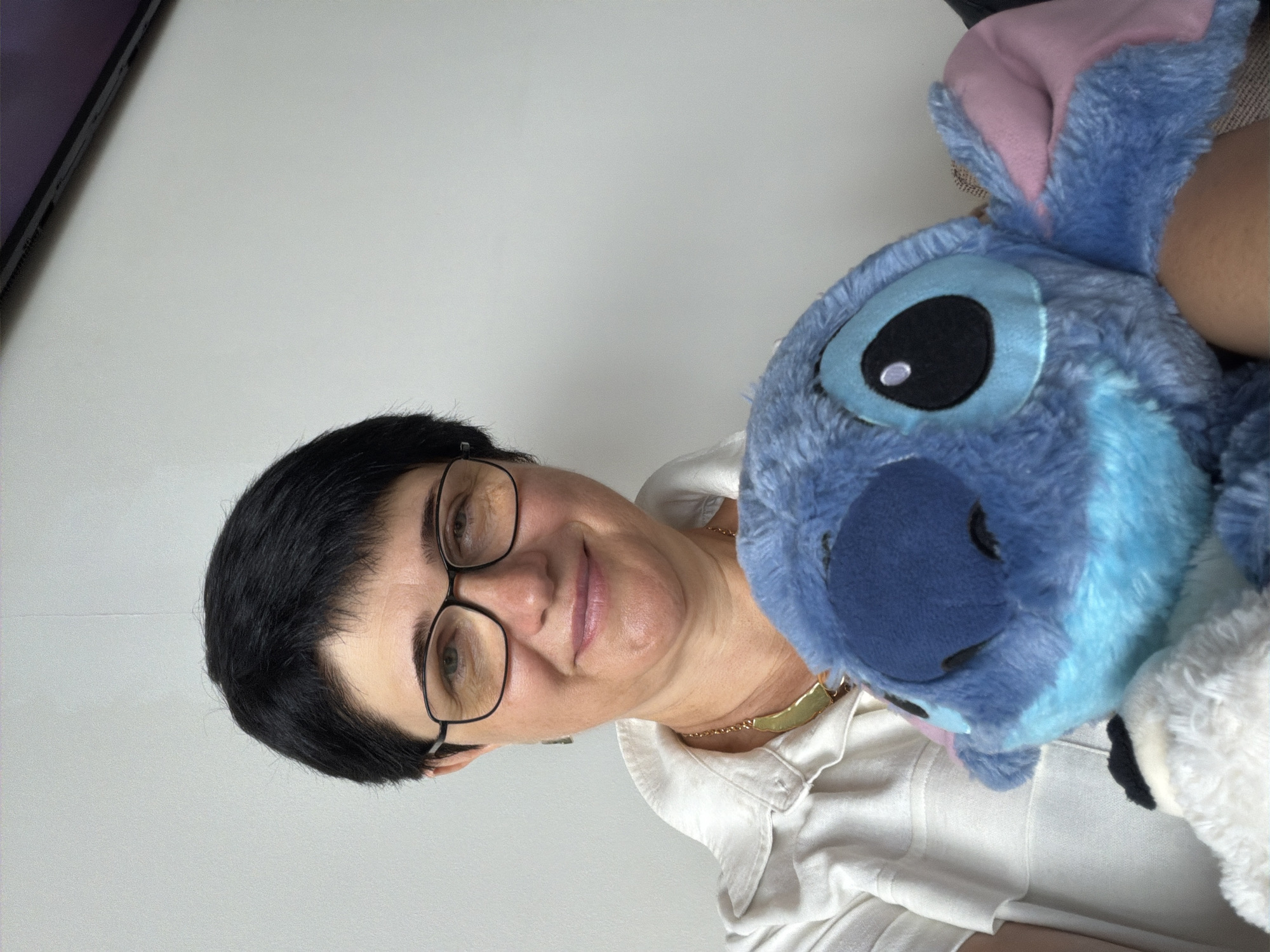 A woman with short black hair and glasses is holding a blue stuffed Stitch toy in front of her. She is wearing a white collared shirt and a necklace. The background is a plain white wall. The image is a selfie, with the woman looking at the camera and smiling.