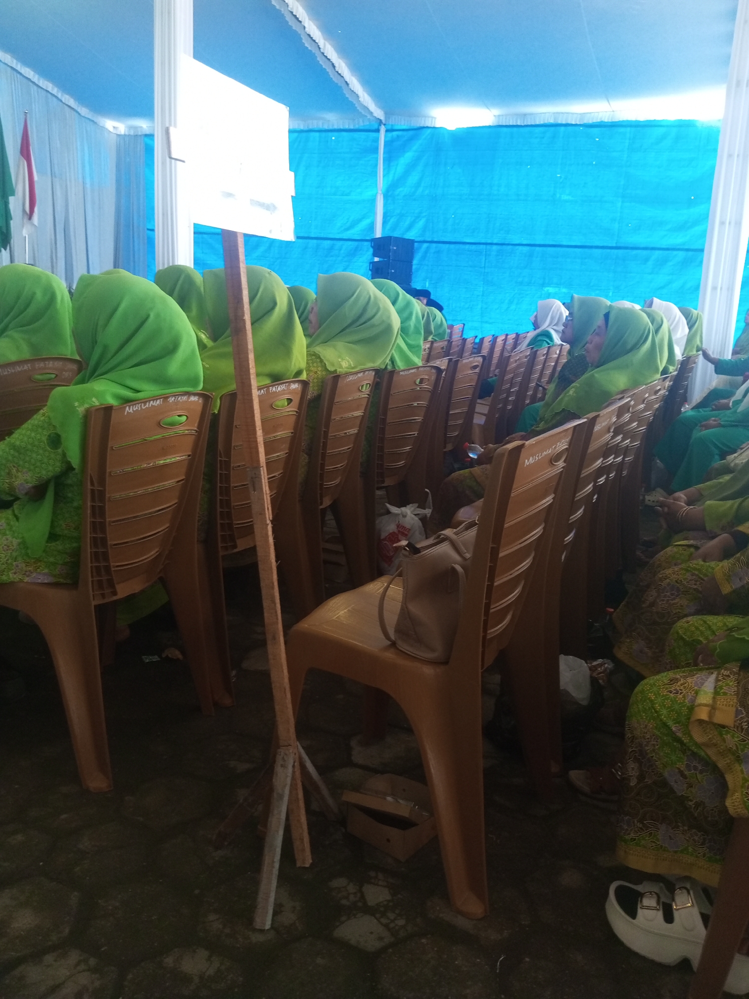 In Klaseman, Indonesia, a group of women are seated on brown plastic chairs arranged in rows. They are all wearing green hijabs and clothing, with some also wearing green dresses patterned with yellow and purple. A white sign on a wooden stand is positioned in the foreground. The scene appears to be outdoors, under a blue tent. The ground is paved with dark hexagonal tiles. Some women are looking forward, while others are looking to the side. There are bags and a water bottle on the floor near the chairs.