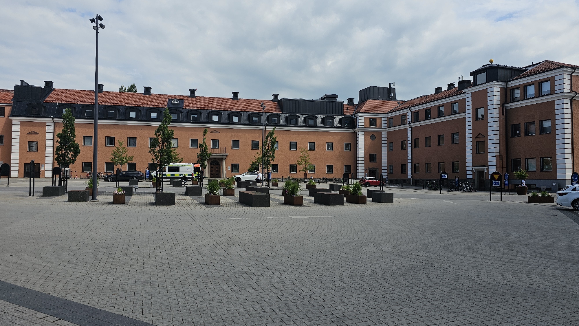 The image depicts a large, brick building with a red tiled roof, located in Linköping, Sweden. The building appears to be a government or administrative facility, with multiple windows and entrances. In the foreground, there is a paved courtyard with several planter boxes and seating areas. A few cars and bicycles are visible in the background, as well as a few people. The sky is overcast, suggesting a cloudy day. The architecture of the building is traditional, with symmetrical elements and decorative trim. The overall scene conveys a sense of order and public space.