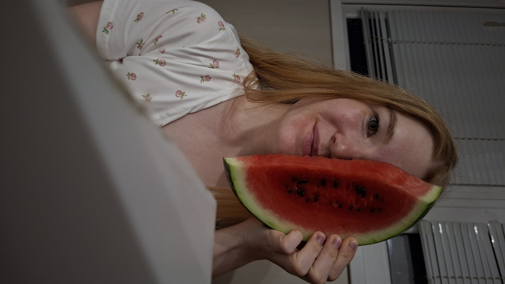 In the foreground of the image, a young woman with long, light brown hair is partially obscuring her face with a slice of watermelon. She is wearing a white t-shirt with a subtle floral pattern. Her left eye and part of her nose and mouth are visible. The watermelon slice is vibrant red with black seeds. The background appears to be an indoor setting, with blinds visible on a window. The lighting is dim, creating a moody atmosphere. The image does not contain any information that would allow for its location in the city of Chaikovsky, Russia.