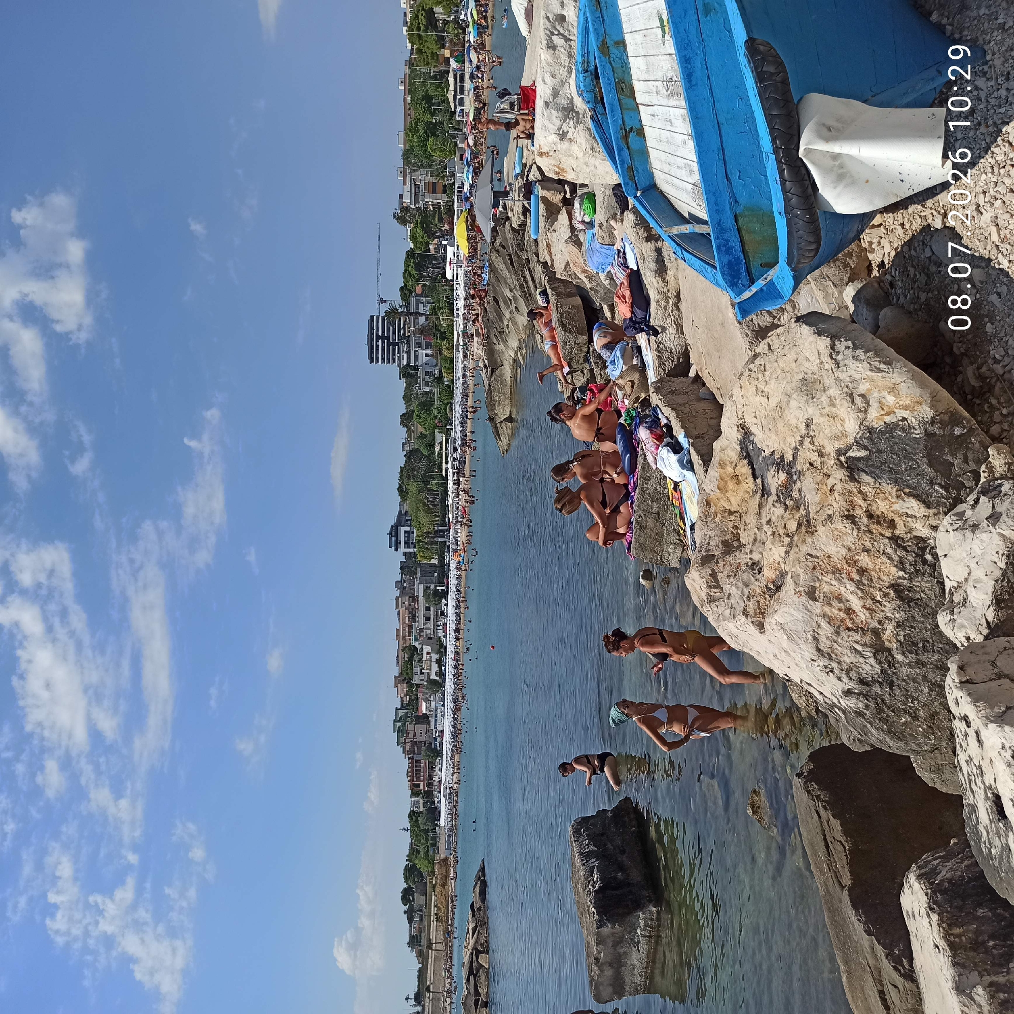 The image depicts a crowded beach in Trani, Italy, on July 8, 2026, at 10:29 AM. The scene captures various individuals engaged in typical beach activities. In the foreground, several people are visible near large rocks and in the shallow water, some are conversing, while others appear to be sunbathing on the rocks or wading. A blue boat, partially visible on the right side of the frame, rests on the shore. The background reveals a densely populated beach with numerous umbrellas and people enjoying the water and sand. Beyond the beach, a cityscape with buildings and trees is visible, characteristic of Trani. The sky is clear with scattered clouds. The presence of many people suggests a popular and lively beach destination.