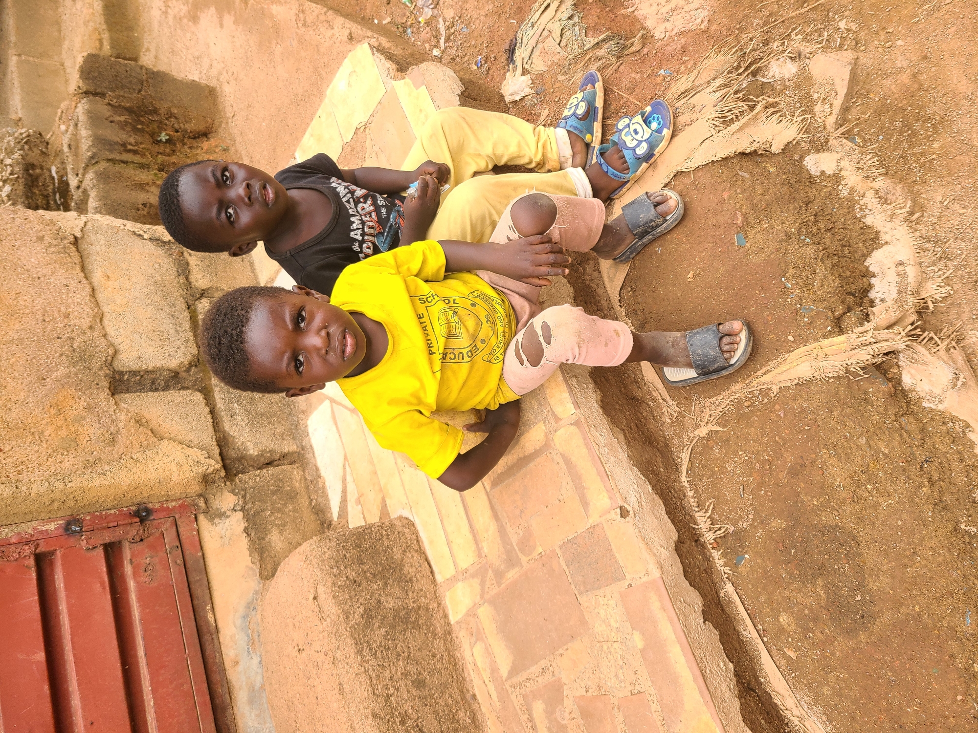 Two young children are seated on a raised brick and concrete surface outdoors on a bright, sunny day, consistent with the location context of Jos, Nigeria. Both children have dark skin and short, dark hair, and are looking directly at the camera with neutral expressions.
The child in the foreground wears a bright yellow short-sleeved t-shirt and light pink or faded orange pants with visible tears at the knees. He has dark sandals on his feet. The yellow t-shirt features text that reads "PRIVATE SCHOL" (school), "EDUCA" (education), "TO EXCEL IS," "OF KNOWLED" (knowledge), and "AND SUCCEED," suggesting it might be part of a school uniform or promotional attire.
Behind him, another child wears a black short-sleeved t-shirt, yellow long pants, and blue sandals adorned with cartoon-like figures. The black t-shirt displays partially legible text including "Je AMAZING DIDER" and "THE SP."
The setting is characterized by reddish-brown bricks and rough concrete structures where the children are sitting. The ground immediately surrounding them is composed of dry, reddish-brown earth and dirt, with some scattered dry organic material. To the far left, a reddish-brown, horizontally ribbed metal panel or door is partially visible. The overall scene suggests a somewhat rustic or undeveloped outdoor environment.