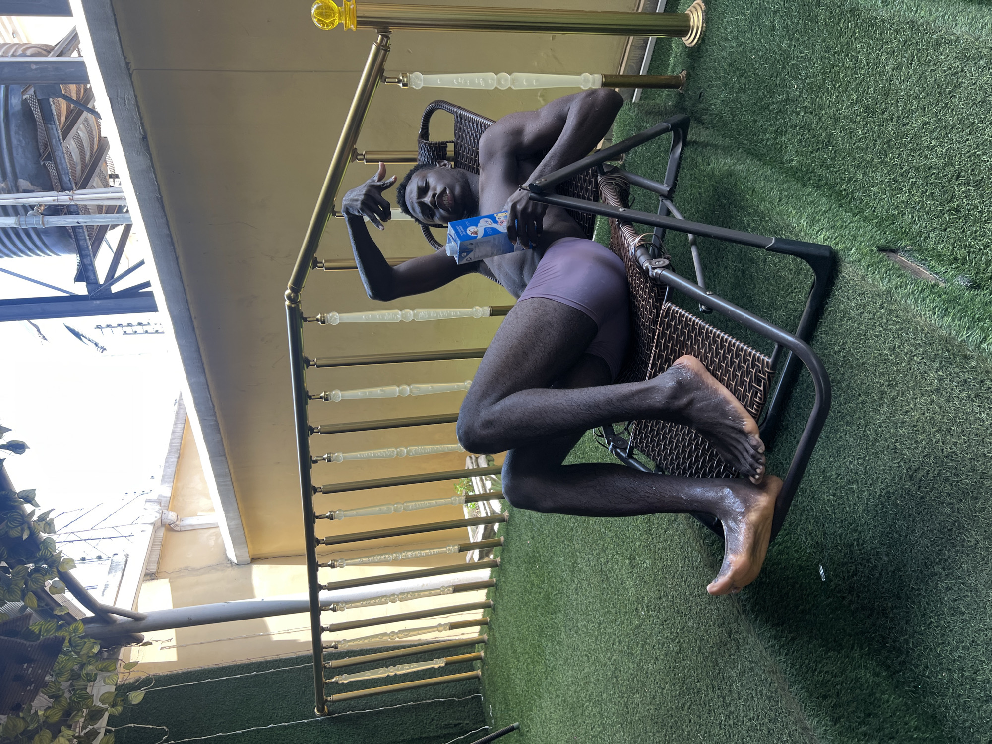 A dark-skinned male is posed in a chair outdoors on a green astroturf surface. He wears purple underwear and holds a blue milk carton. He makes a thumbs up gesture with his right hand. A gold and clear balustrade is partially visible in the background, and a building is seen above it. The location is presumed to be in an unidentified area in an unknown city.