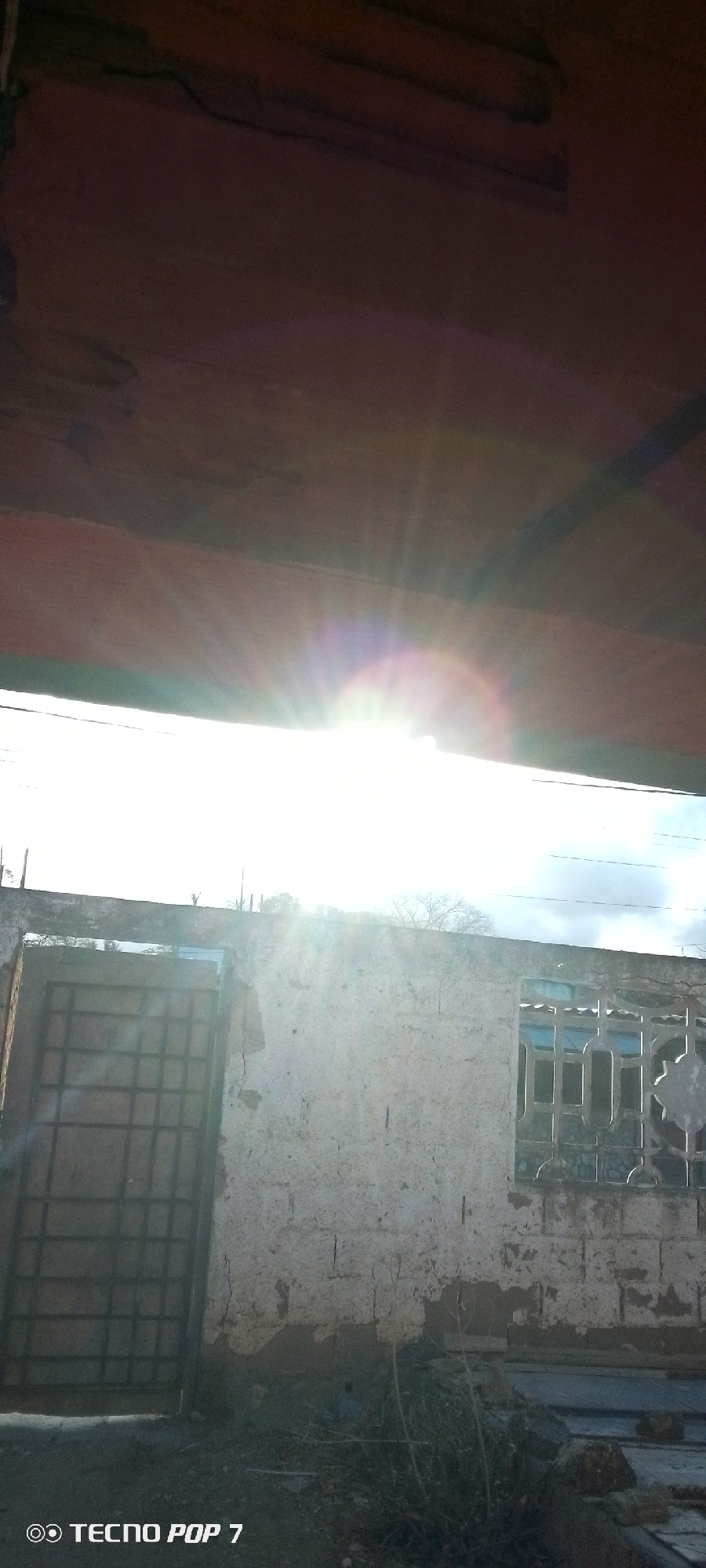 The image shows a dilapidated building with a partially collapsed roof in Altagracia, Venezuela. The sun shines brightly through the gap in the roof, creating a glare. The building appears to be constructed of concrete blocks, with patches of crumbling plaster. There is a barred window on the right side of the wall, and a barred doorway to the left. The ground in front of the building is cluttered with debris and vegetation. There are power lines in the sky.