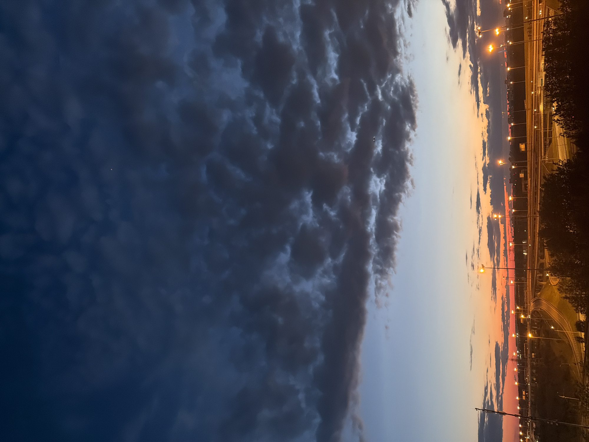 The image depicts a twilight scene over a city, likely at dusk or dawn. The sky is a dramatic gradient of deep blue and charcoal gray clouds in the upper portion, transitioning to hues of orange, pink, and pale blue closer to the horizon. A few scattered clouds are visible in the upper sky, one of which appears to have a faint, distant star or light source.
Below the sky, a complex network of elevated highways and roads dominates the midground and foreground. Numerous streetlights are illuminated, casting a warm glow across the asphalt and surrounding vegetation. The city skyline is visible in the distance, a blur of structures and lights against the horizon. Trees and foliage are present in the lower foreground, silhouetted against the brighter elements of the scene. There are no discernible people or specific actions captured in the image; it is a static view of an urban landscape at a transitional time of day. The overall impression is one of urban infrastructure illuminated by artificial light under a moody sky.