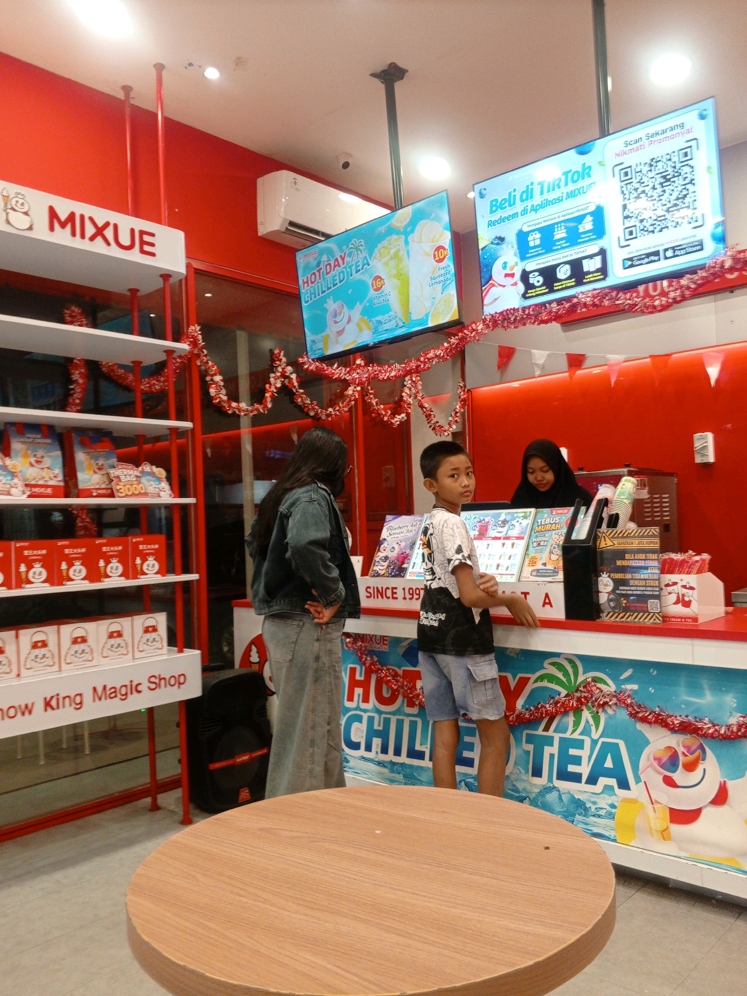 A medium shot captures a Mixue store in Gabus, Indonesia. A young boy in a black and white t-shirt and denim shorts stands at the counter, looking towards the viewer. Behind the counter, a woman wearing a black hijab serves drinks. The store is decorated with red and white festive garlands. Large digital screens display advertisements for Mixue products. Shelves on the left are stocked with merchandise, including bags and boxes featuring the Mixue mascot. A round wooden table is visible in the foreground. The environment is brightly lit, with red walls and a white ceiling.