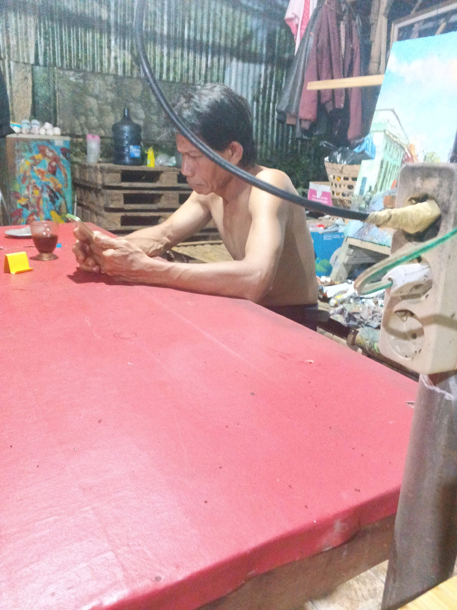The image shows a man sitting at a red table, holding a mobile phone. He is shirtless and appears to be focused on the phone. Behind him, there is a cluttered background with various items, including what looks like a painting, a water dispenser, and stacked wooden pallets. The setting appears to be an outdoor or semi-outdoor space, possibly a workshop or a backyard, with a corrugated metal wall in the background. A black cable is prominently visible, arching over the man's head. The overall impression is that of a candid moment captured in an informal environment. The specific location in Ungaran, Indonesia, is not evident from the image itself.