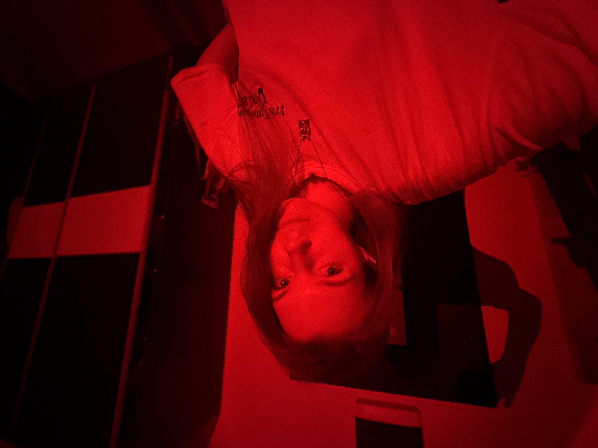 An inverted portrait shows a young woman looking directly at the camera, bathed in intense red light. Her head is positioned towards the bottom of the frame, with her body extending upwards (in the original, inverted orientation). Her long, dark hair frames her face and falls around her. She wears a light-colored, long-sleeved shirt.
On her shirt, near the neckline, the text "It's Time" is visible, along with another word that appears to be "Verfa," both in reverse due to the image's inversion. A small, rectangular pendant hangs from a necklace on her chest.
The background is dark, likely an indoor setting, featuring dark panels or walls with prominent red-lit horizontal and vertical strips or structures. The strong red light creates dramatic contrasts and deep shadows throughout the scene. A shadow, possibly of an arm or another figure, is cast on a red-lit surface to the right. The overall atmosphere is stark and moody, defined by the monochrome red illumination. The location is Vozdovac Urban Municipality, Serbia.