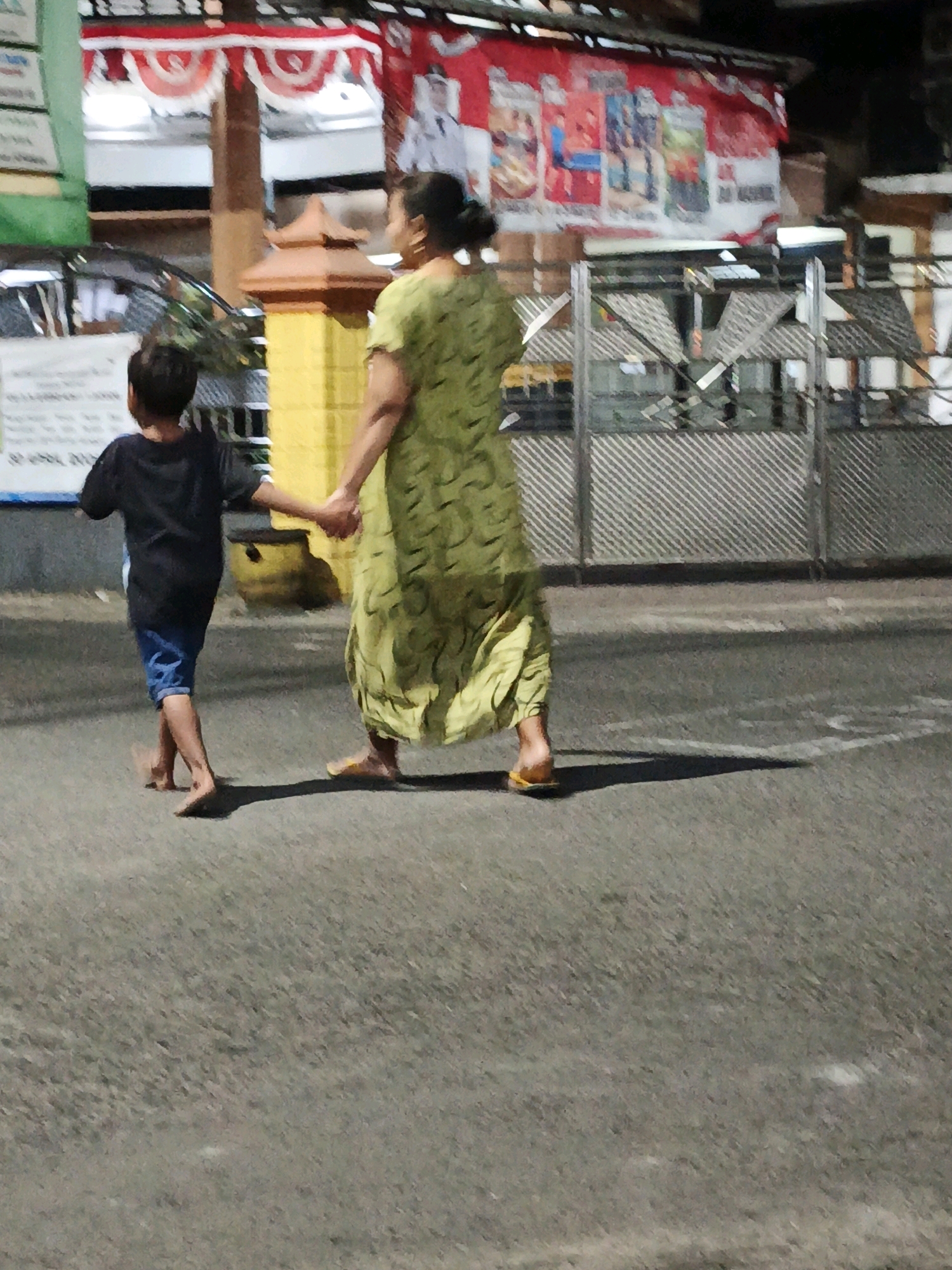 In Ketegan, Indonesia, a woman and a young boy are walking hand-in-hand on a paved street at night. The woman, wearing a long, light green patterned dress, is barefoot with yellow sandals on her feet. The boy, dressed in a black t-shirt and denim shorts, is also barefoot. They are walking away from the viewer, towards a decorative gate and a building with banners displaying advertisements. The background shows a street scene with various structures and signage illuminated by artificial light.